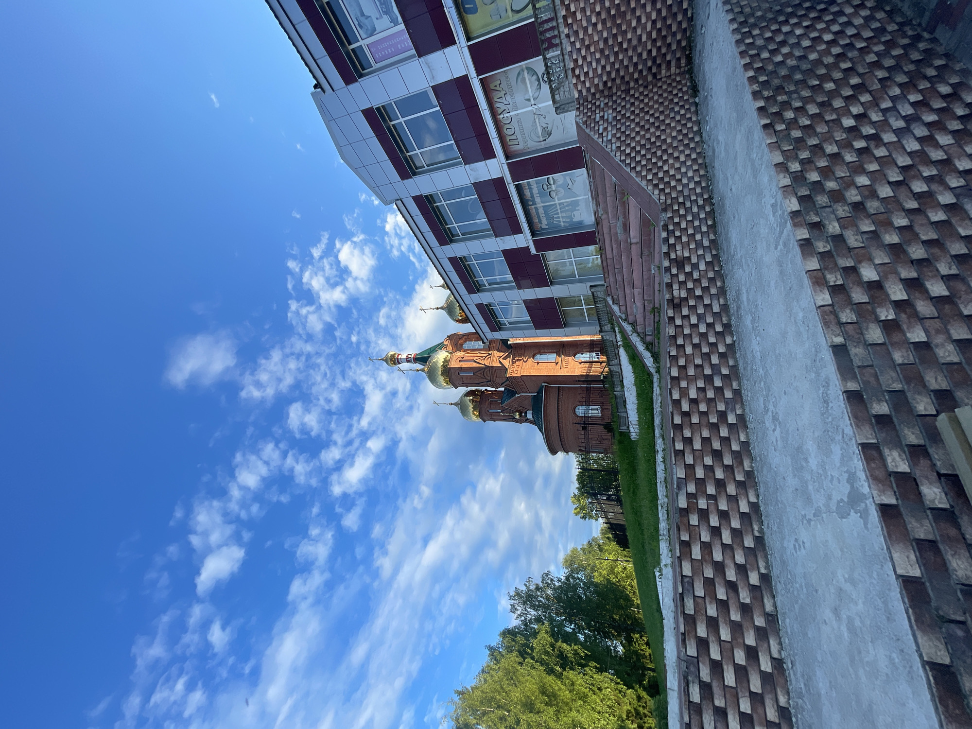 The image shows the Church of St. Sergius of Radonezh and a modern commercial building, both located in the settlement of Krasnye Baki, Russia. The church is a red brick structure with golden domes, situated on a grassy hill. Adjacent to the church is a multi-story building with a facade of silver and burgundy panels, featuring large windows. In the foreground, there are concrete stairs and retaining walls constructed with patterned brickwork. The sky is blue with scattered white clouds. There are no people visible in the image.