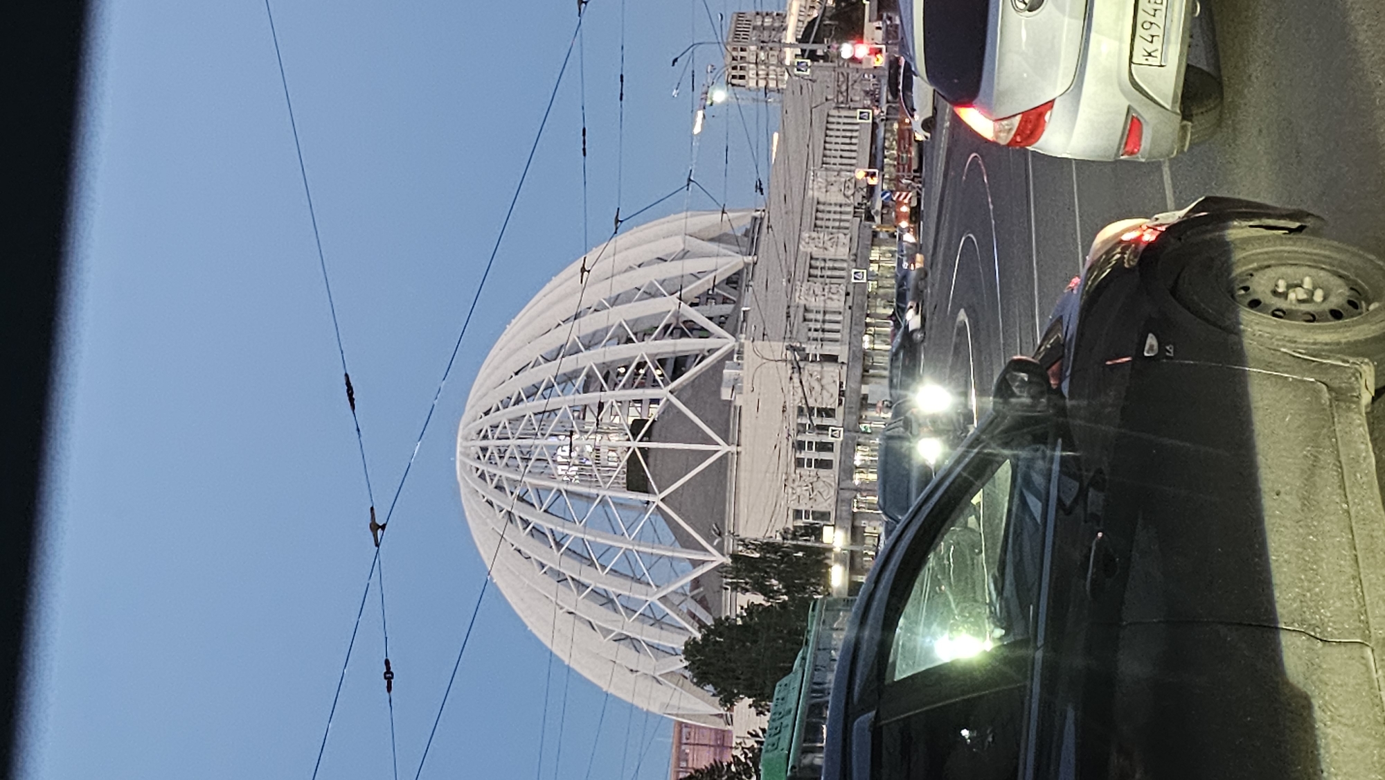 The image shows a view of the Yekaterinburg Circus in Yekaterinburg, Russia, from a street level perspective. The iconic geodesic dome of the circus building is prominent in the background, its white lattice structure clearly visible against a pale blue twilight sky. The building's facade features decorative elements and windows. In the foreground, there is a street with several cars, suggesting a traffic scene. A dark-colored car is positioned in the lower left of the frame, partially obscuring the view of other vehicles and the road. A silver car is visible to the right, with its license plate showing "K 494B". Overhead wires are present in the sky, likely for trolleybuses or trams, and streetlights are visible in the distance. The overall scene depicts an urban environment during the evening or early morning hours.