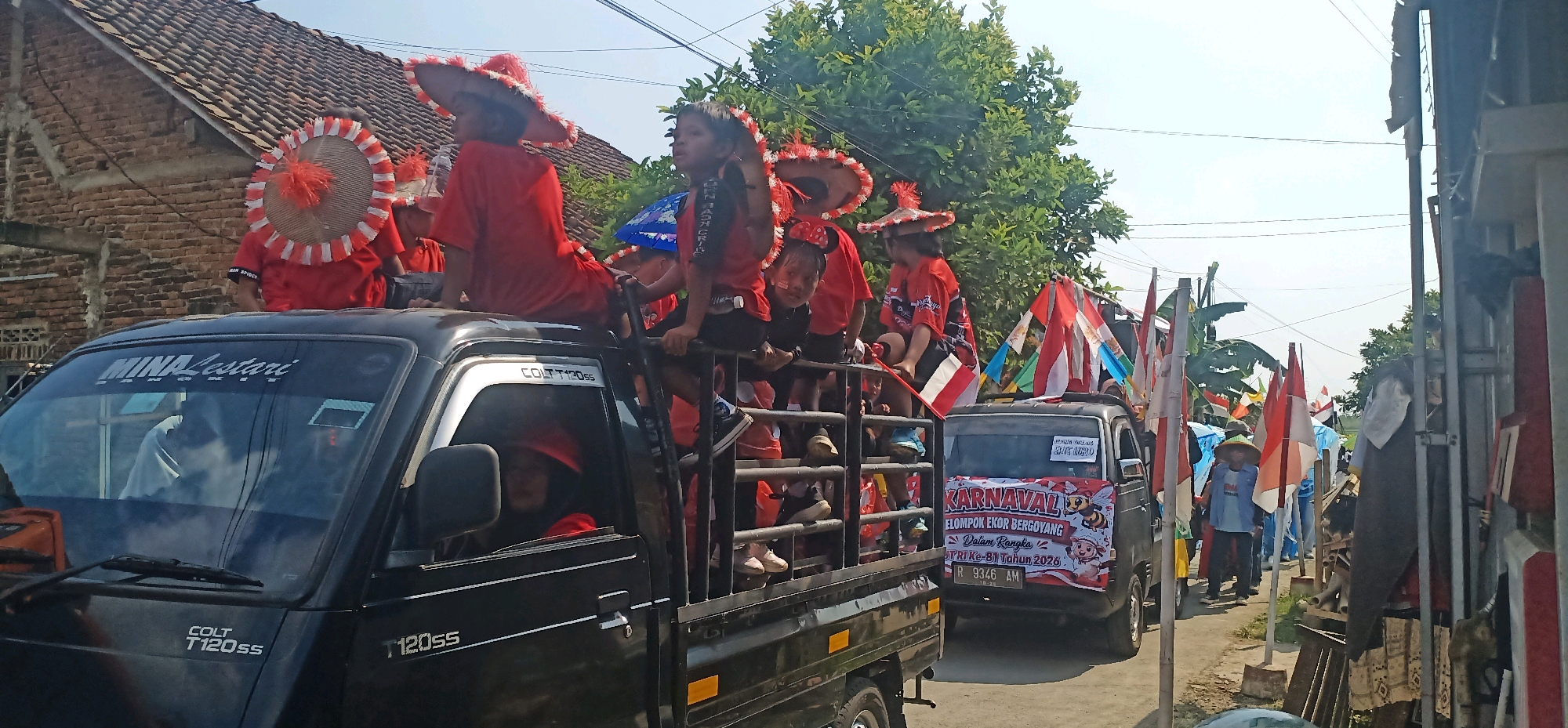 The image depicts a parade in Kincang, Indonesia. A black pickup truck, identified as a Colt T120ss, is prominent, carrying several children dressed in red attire and festive hats adorned with white and red fringe and orange pom-poms. Some of the children are also wearing red shirts with white lettering. The truck has a banner on its rear that reads "KARNAVAL" in large letters, along with smaller text and a cartoon bee graphic. Another truck is partially visible behind the first, also adorned with flags. Indonesian flags, white and red, are displayed throughout the scene, some on poles and others attached to vehicles. The parade appears to be moving down a street lined with buildings and trees. Several people, including adults, are seen on the side of the street, observing the procession. The sky is clear and bright, suggesting a sunny day. The overall atmosphere suggests a community celebration or event.