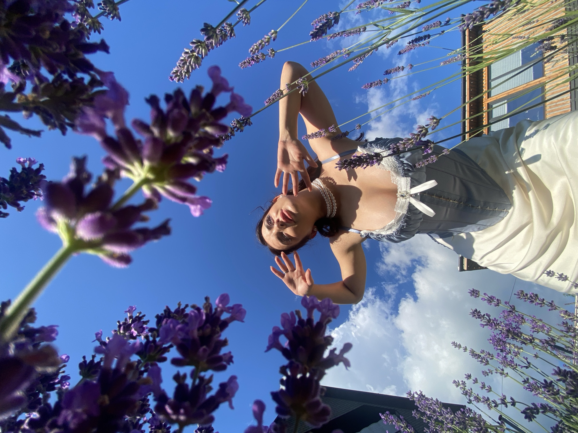 A low-angle shot captures a person among lavender flowers, with a bright blue sky and white clouds overhead. The person, wearing a light-colored satin dress and a pearl necklace, is upside down in the frame, their head and torso tilted downwards. They are reaching out with one hand, fingers splayed. The lavender flowers are in focus in the foreground and scattered throughout the image, their purple blooms contrasting with the blue sky. In the background, a building with wooden siding and windows is visible. The lighting suggests it is daytime, likely sunny. The overall mood is whimsical and serene. No text is visible.