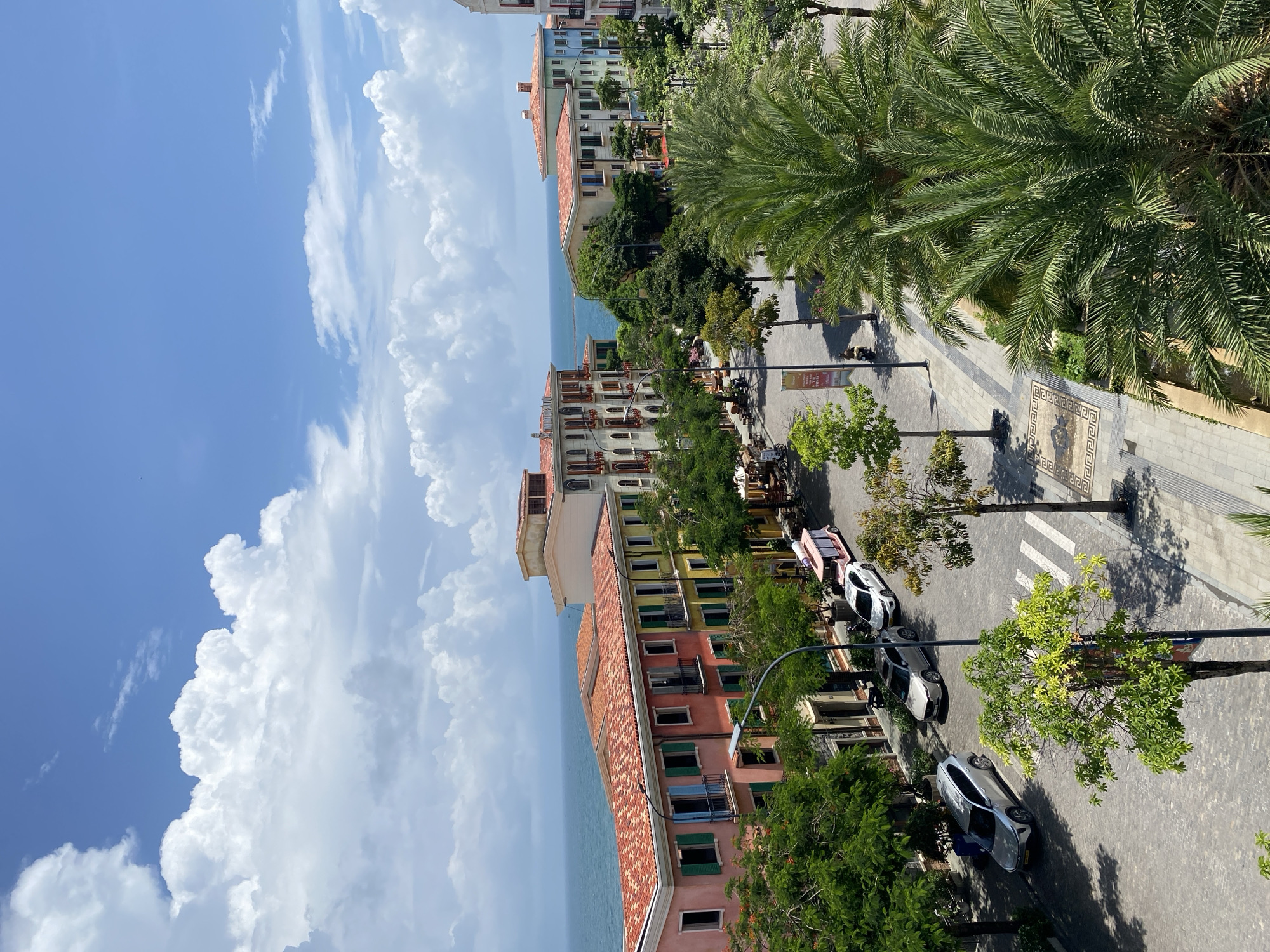 The image shows a bright, sunny day in Phú Quốc, Vietnam, specifically a street scene with colorful buildings reminiscent of Mediterranean architecture. The buildings, painted in vibrant hues of pink, orange, yellow, and blue, have terracotta tile roofs and are situated along a street lined with lush green trees, including palm trees. The street itself is paved with cobblestones and features a pedestrian walkway.
Several cars are parked along the street, including a silver SUV, a white sedan, and a small pink and white vehicle that appears to be a golf cart or small shuttle. People are visible in the distance, one riding a scooter. The street leads towards a body of water, likely the ocean, which is visible in the background under a bright blue sky filled with fluffy white clouds. The overall impression is of a resort town or a well-maintained urban area with a strong European influence.