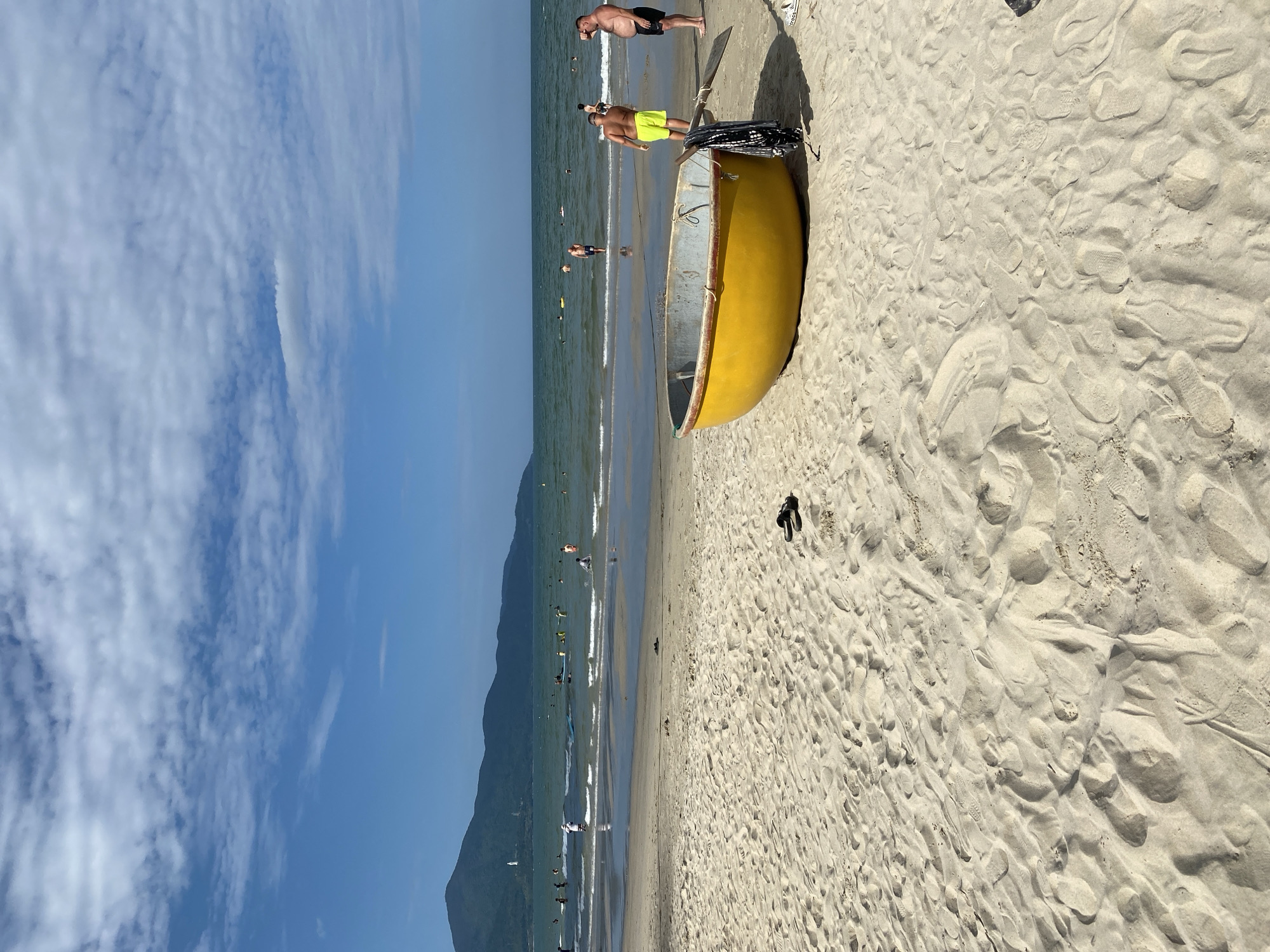 This is a photograph of My Khe Beach in Da Nang, Vietnam, taken on a sunny day. The foreground is dominated by rippled sand, showing numerous footprints and a pair of black sandals near the upper right. In the mid-ground, a bright yellow, round basket boat, commonly used by local fishermen, rests on the sand, with a black and white patterned cloth and a wooden oar leaning against it. Two men are standing near the boat. The man on the left wears yellow swim shorts, and the man on the right wears black swim shorts. The sea is visible in the background, with small waves breaking on the shore. Several people are in the water, some swimming and others standing. Further in the distance, a mountain range forms the horizon under a blue sky with scattered white clouds.