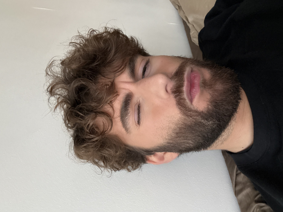 A close-up, chest-up portrait shows an adult male subject with brown, curly hair and a full brown beard. His complexion is fair, with a subtle rosy tone on his cheeks. His right eye is completely closed, while his left eye is partially squinted. His eyebrows are furrowed, creating lines on his forehead, and his mouth is slightly pursed, conveying an expression of confusion, discomfort, or intense thought. The subject is wearing a plain black crew-neck shirt or sweatshirt. The background consists of a uniformly light-colored, possibly white or off-white, wall to the left and top of the subject, with a soft, light brown or beige object, likely a cushion or bedding, visible on the lower right, suggesting an indoor setting. The precise location within the city of Paris, France, cannot be determined from the visual evidence provided in the image.