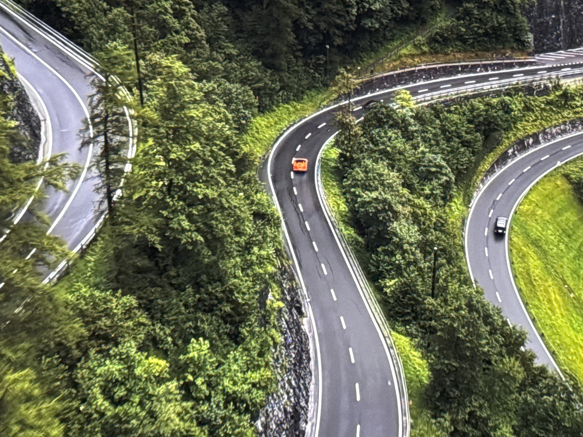 An aerial, high-angle, close-up shot captures a winding, multi-lane asphalt road traversing a lush, verdant mountainside. The road features white lane markings and appears to be wet, reflecting the surrounding greenery. Two vehicles are visible: an orange sports car is navigating a tight curve, while a dark-colored sedan is further down the road on a wider bend. The environment is dominated by dense, dark green trees and foliage, with patches of bright green grass on the slopes. The scene is devoid of any urban structures, suggesting a rural or mountainous setting. There is no indication of the city of Burgas, Bulgaria, in this image.