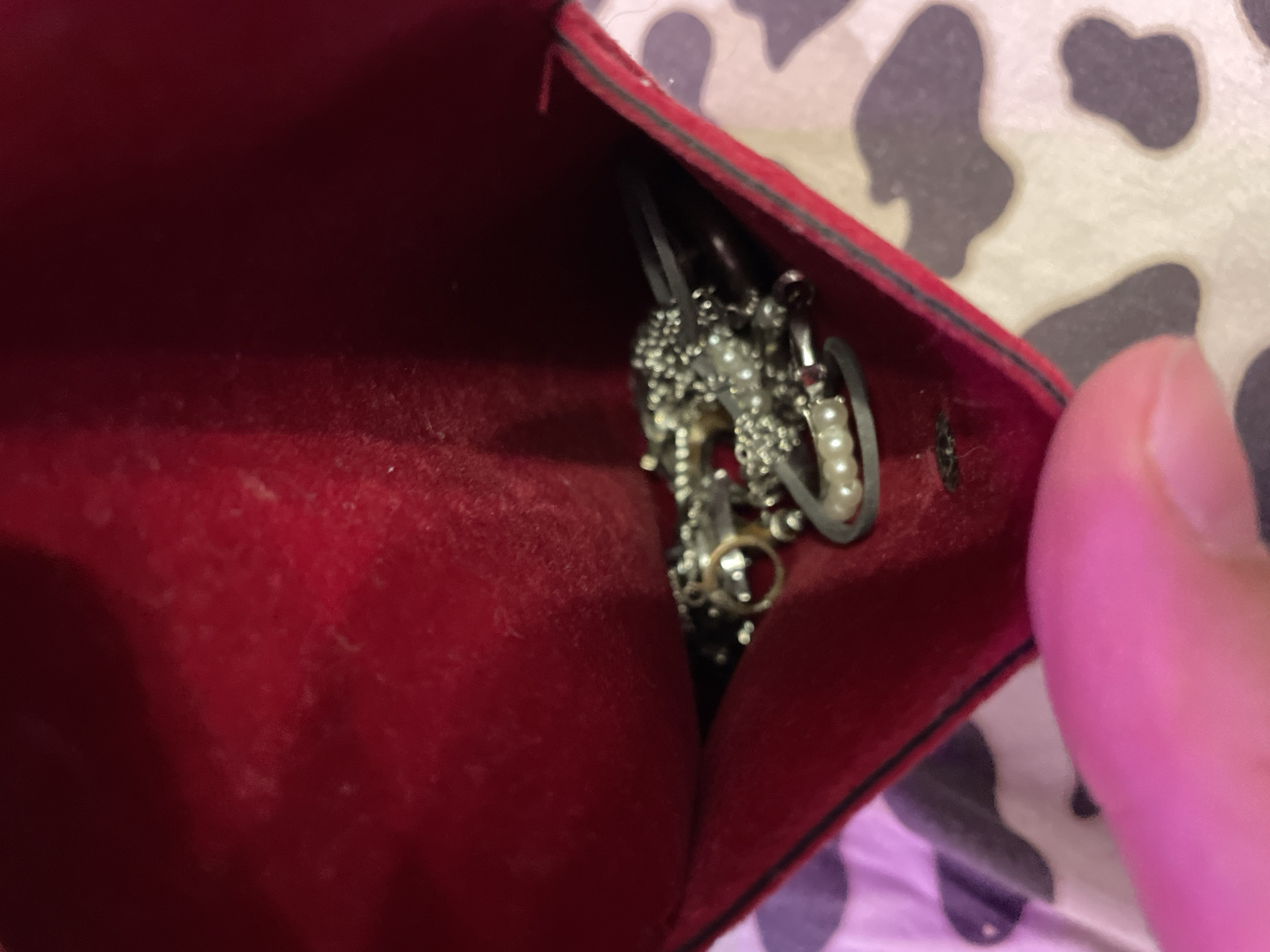 A close-up, low-angle shot shows a person's thumb and forefinger gently holding open a small, red velvet jewelry pouch. Inside the pouch, a collection of silver-toned jewelry is visible. This includes several rings, one with a small red gem, and a bracelet adorned with small pearls. The jewelry is clustered together within the pouch. The background consists of a patterned surface, possibly a bedsheet, with a cow-print-like design in shades of beige and grey. The overall scene is dimly lit with a pinkish hue, likely from an artificial light source. The location is not discernible from the image.