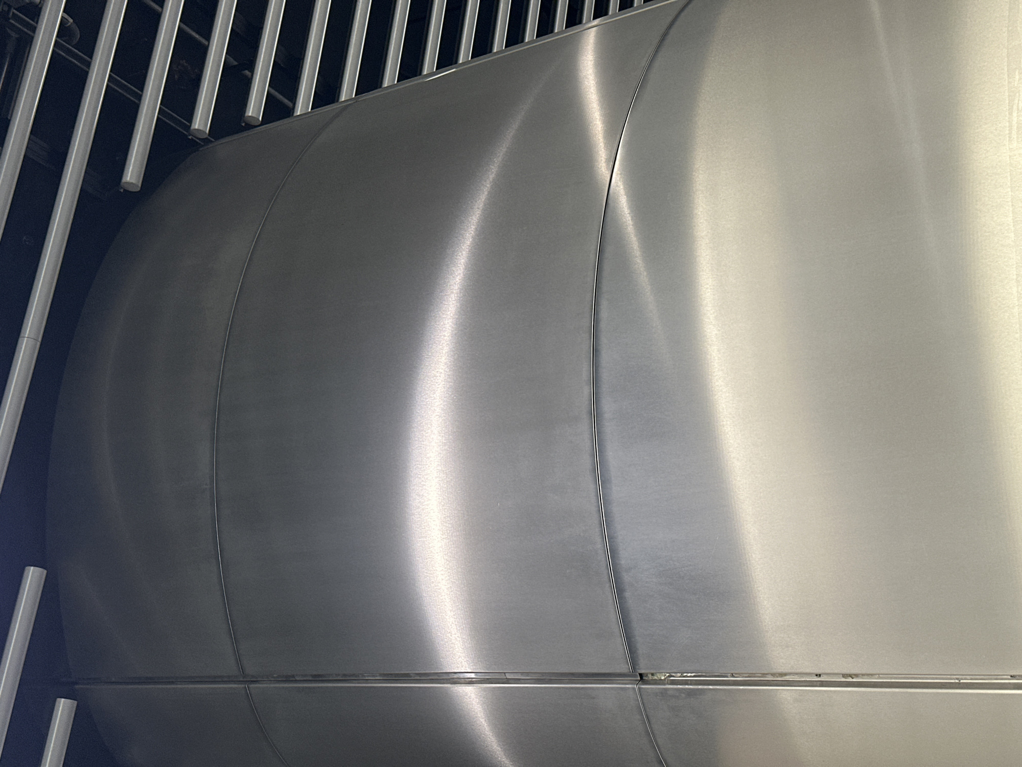 The image shows a large, horizontal, cylindrical metal tank, possibly made of stainless steel, situated within an enclosed space defined by a series of vertical, linear metal elements. The tank appears to be composed of multiple sections joined together, indicated by the vertical seams. The surface exhibits variations in reflective intensity due to the curvature and lighting conditions. The vertical metal elements in the surrounding space are evenly spaced and suggest some form of enclosure or structure related to the tank. The environment is indoors and appears to be industrial or utility-related, given the size and type of tank, and the bare metal construction. There is no indication of the city of London, United Kingdom from this image. There are no people present.