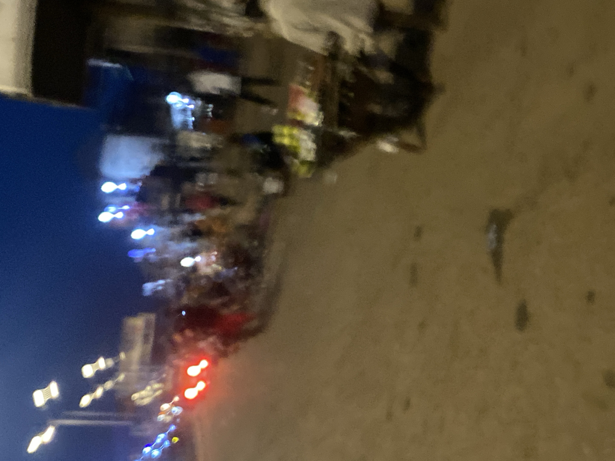 The image depicts a blurry, night-time street scene in Bukuru, Nigeria. Several streetlights are visible, casting a dim glow on the unpaved road. In the foreground, a street vendor's cart is laden with yellow objects, possibly fruits or bottles, and other indistinct items. Several people are present, but their features are obscured due to the low light and blurriness. Some appear to be walking, while others are gathered around the vendor's cart. The overall impression is one of a bustling, albeit somewhat chaotic, urban environment at night.