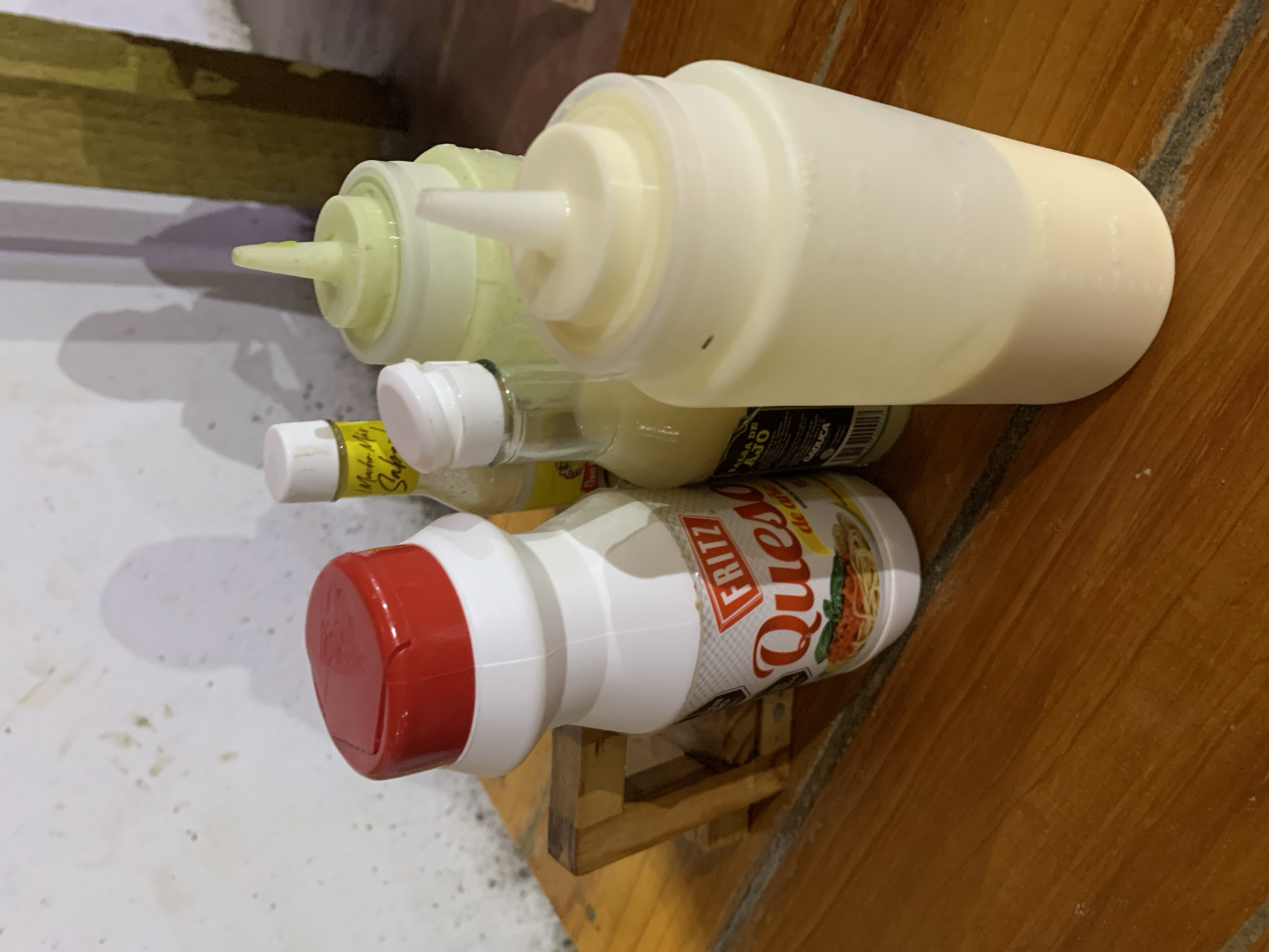 This image captures a close-up, angled view of several condiment bottles resting on a wooden surface, likely a table or shelf, against a light-colored, possibly textured wall or floor in the background. The scene suggests an indoor setting, possibly a kitchen or dining area, illuminated by artificial light which casts visible shadows.
The primary subjects are four bottles of various sizes and types. On the far left, partially obscured, is a large, translucent squeezable bottle with a white cap, containing a pale greenish-yellow liquid. Next to it, another similar large squeezable bottle with a white cap holds a creamy white substance, possibly mayonnaise or a white sauce. A small black label on this bottle is partially visible, showing text that includes "SA..." (likely part of "SALSA") and "GAOLICA," along with a barcode.
Below these larger bottles is a smaller, clear glass bottle with a white cap and a yellow label, containing a light-colored liquid. Text on its label clearly reads "Salsa de Ajo" (Garlic Sauce), indicating its content. Further to the right, a white plastic bottle with a distinctive red flip-top cap stands on a small wooden support structure. This bottle features a colorful label with the brand "FRITZ" and "Queso" (Cheese) prominently displayed, alongside an illustration of pasta with sauce, suggesting it contains a cheese-flavored sauce.
No human subjects are present, and there is no discernible activity taking place, rather a display or storage arrangement of these items. The presence of "Salsa de Ajo" and "Fritz Queso" brands, combined with the provided location context of Santa Ana, Venezuela, points to a Latin American culinary setting. The time of day is not clear, but the evident shadows and indoor environment imply artificial lighting.