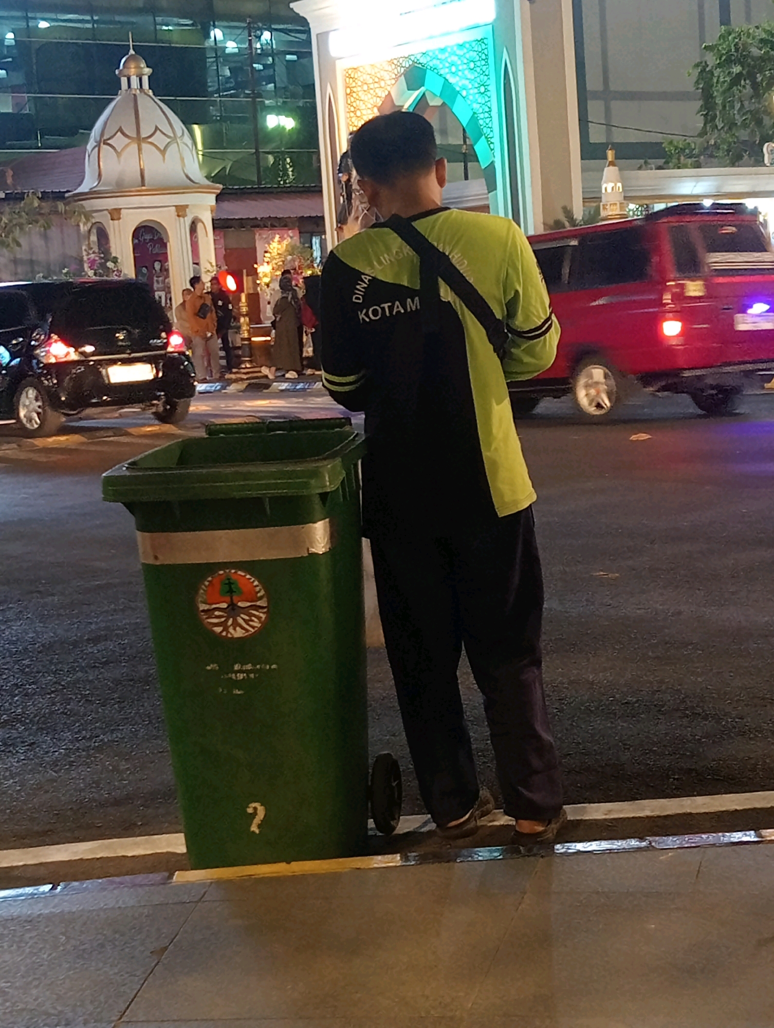A man wearing a neon green and black uniform, possibly a sanitation worker, stands on a sidewalk next to a green trash bin. The trash bin has a circular emblem on it and a white question mark painted on its side. The man is facing away from the camera, looking towards a street scene. In the background, a red van is driving past, and a black car is parked. There are buildings with illuminated architectural features, suggesting an urban setting at night. The overall impression is a street scene in Madiun, Indonesia, likely near a commercial or public area, with the sanitation worker attending to his duties.