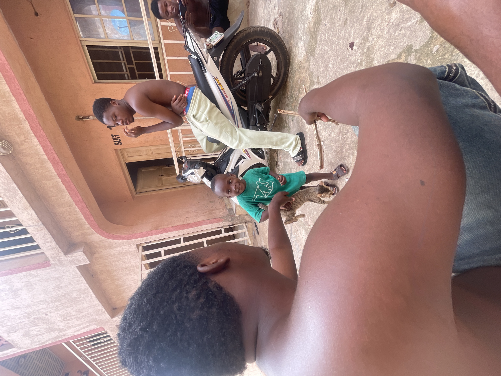 A daytime, wide-angle outdoor shot reveals a group of people in Lokoja, Nigeria. An unidentifiable building stands in the background. In the immediate foreground, a person wearing denim jeans sits with their back to the camera, holding wooden sticks. A child in a green t-shirt stands next to them, petting a tabby cat. Near the child, a man with no shirt leans against a parked white motorcycle. Another person, also shirtless, is seen standing near the motorcycle. The overall setting appears to be a residential area.