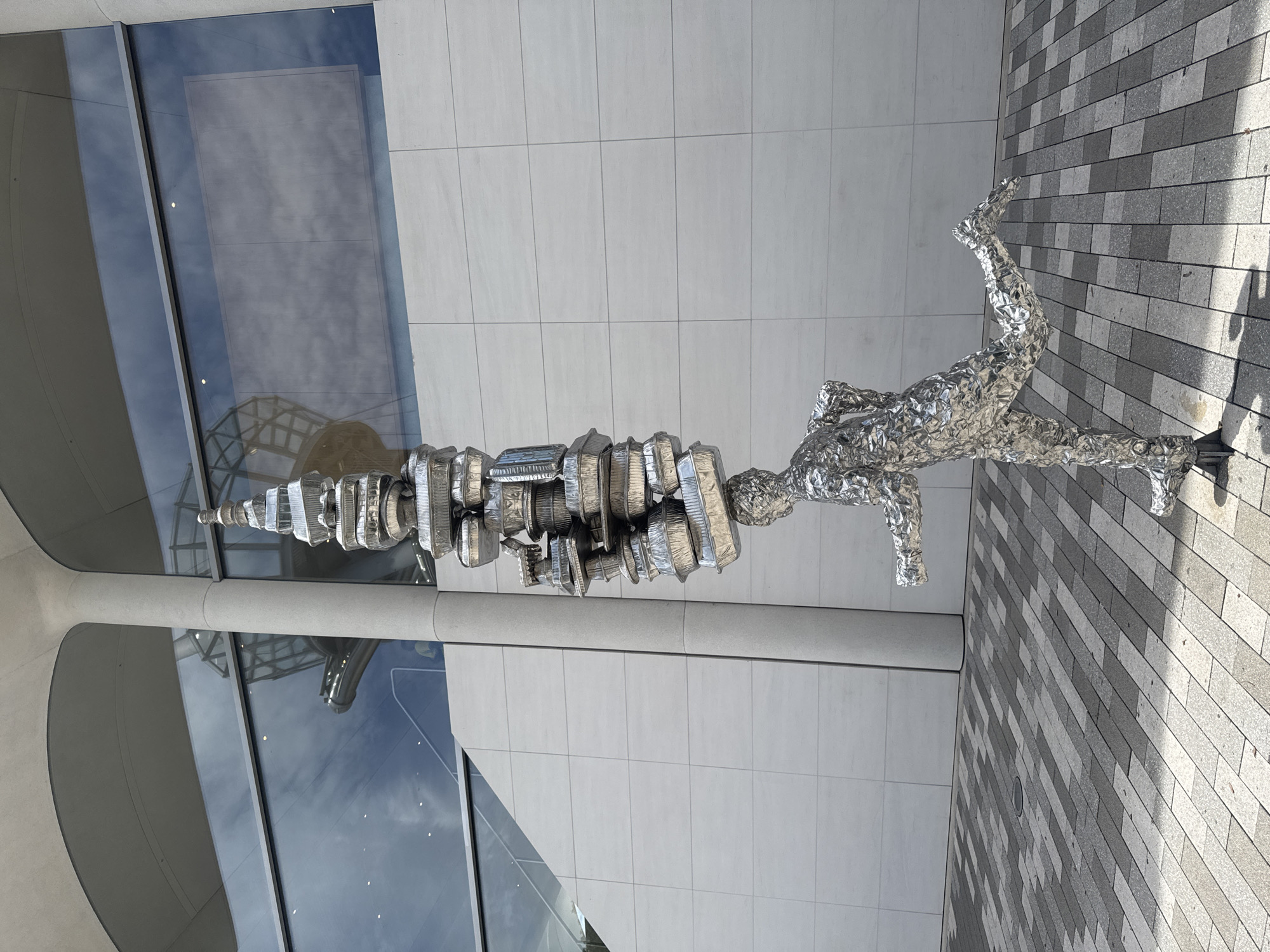 This media file shows a metal sculpture of a person in motion, outdoors. The sculpture is made of what appears to be crumpled aluminum foil, giving it a textured, almost crystalline appearance. The figure is depicted mid-stride, with one leg extended backward and the other bent forward, suggesting a running or dancing pose. The body is elongated and somewhat abstract, with the head and torso forming a single, continuous element that is stacked with various metallic, disc-like shapes.
The setting is a modern urban environment. The sculpture is placed on a walkway paved with rectangular gray and black tiles. Behind the sculpture, there is a building with a facade of large, light-colored rectangular tiles. A large section of the building features a smooth, curved, light gray architectural element. To the left of the sculpture, there is a large glass window that reflects the sky and the surrounding architecture, including a glimpse of a domed or arched structure within the building.
The time of day appears to be daytime, with sunlight casting shadows on the tiled walkway, indicating a bright and possibly sunny day. The shadows are sharp, suggesting the sun is not directly overhead. The overall mood conveyed by the sculpture and its setting is one of dynamism and modern artistry. There is no visible text in the media file. The location is identified as Aventura, United States of America.
