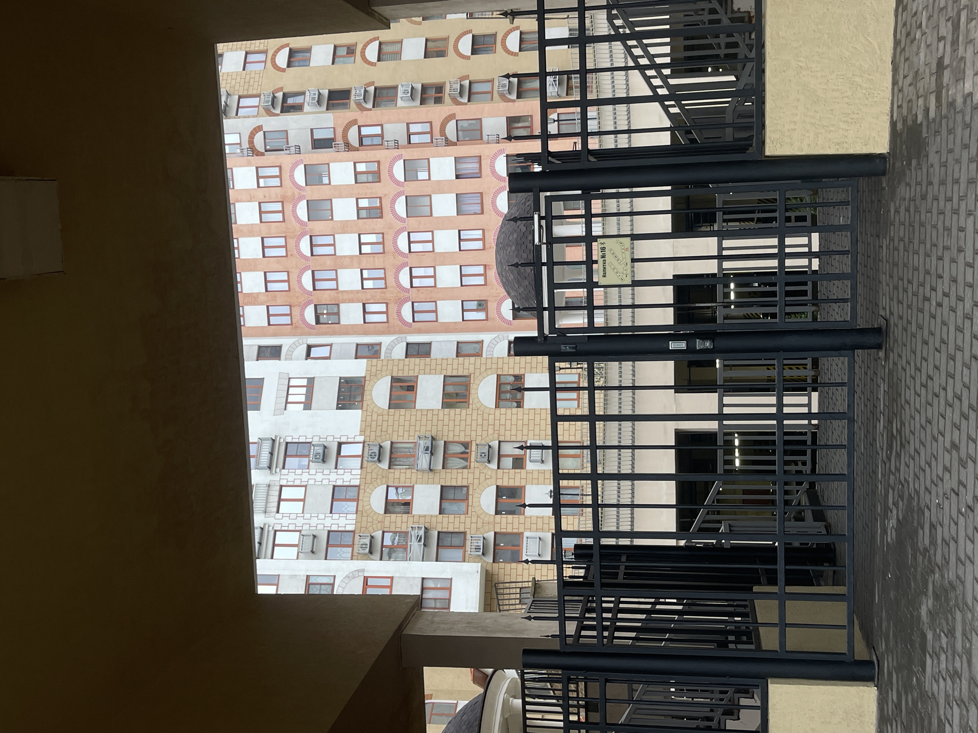 The image captures a view from an elevated position, looking out towards a multi-story building in Razvilka, Russia. The building's facade features a repetitive pattern of windows, some with air conditioning units. The exterior is adorned with a decorative motif of half-circles above the windows. A dark metal staircase and gate structure are in the foreground, partly obstructing the view of the building. The ground is paved with rectangular concrete pavers. The overall environment is urban, characterized by the dense building and constructed hardscape.