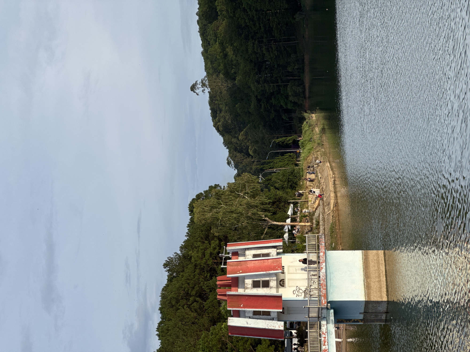 The image depicts a building with a distinct red and white roof structure, situated next to a body of water. Lush green vegetation covers a hillside adjacent to the building. The sky above is overcast. A group of people is visible along the shoreline.
The location is Da Lat, Vietnam.