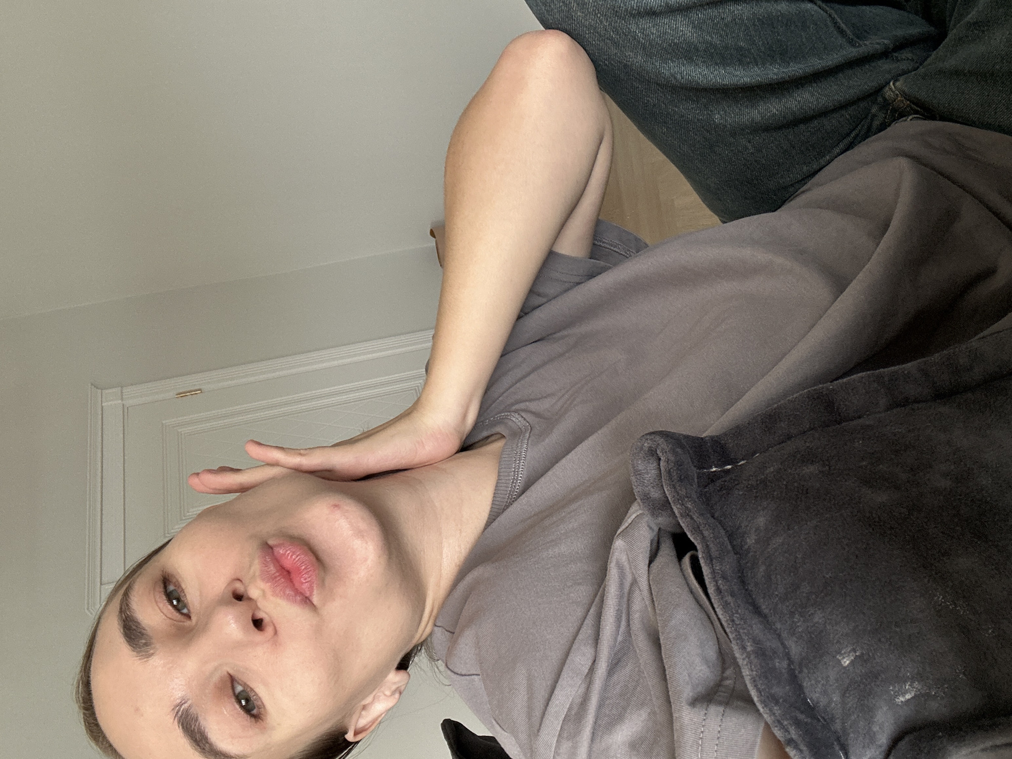 A woman with light skin, brown eyebrows, and green eyes is facing the camera. Her lips are slightly pursed. Her right hand is raised, with the fingers touching her cheek and jawline. She is wearing a gray t-shirt and appears to be sitting. She has a gray blanket on her lap. The background consists of a white wall with a white door and molding. The location is not evident in the image.