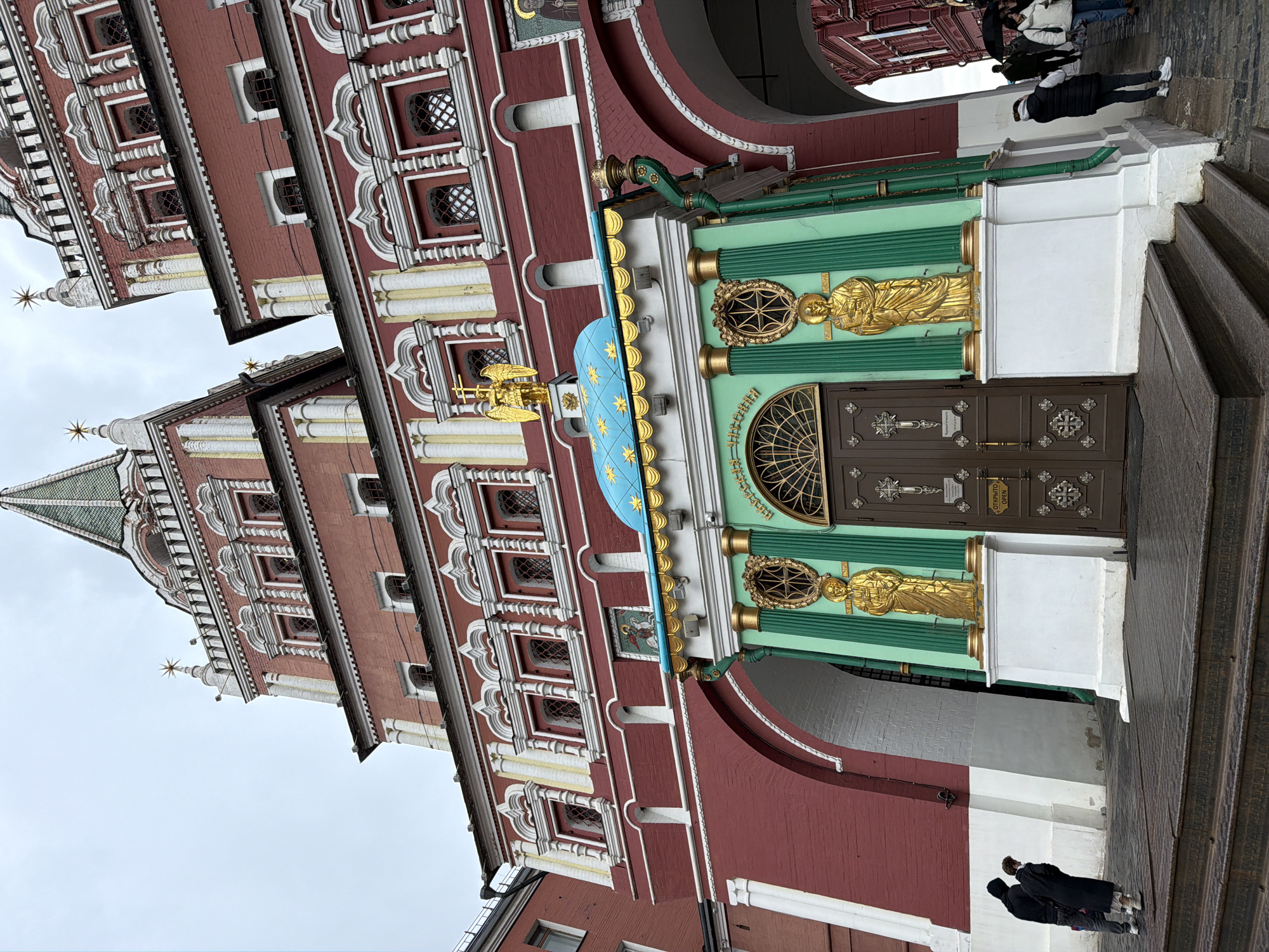 The image is a low-angle shot of an ornately decorated building in Moscow, Russia. The architecture features red brick, white detailing, and multiple tiers with arched windows. A green structure with gold figures and a blue shield adorned with gold stars is attached to the side. A dark brown double door with metal accents is visible within the green structure. A group of people is standing in an archway above the green structure. At the bottom of the image, two figures in dark clothing are visible near the steps leading to the building. The sky above is overcast.