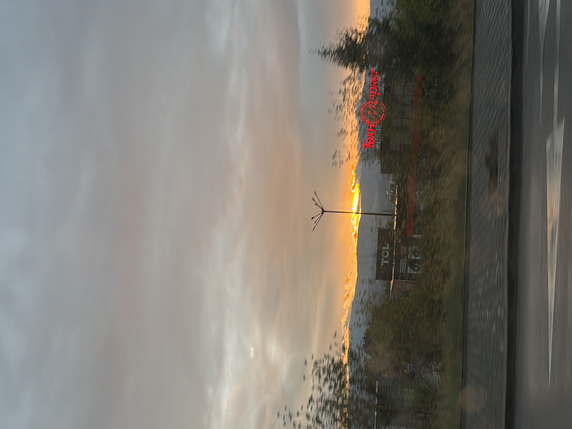 The image depicts a view of the city of Blagoevgrad, Bulgaria, during sunset. The sky is filled with soft, hazy clouds in shades of gray and pale orange, with the setting sun casting a warm glow behind a mountain range. In the foreground, a street or road is visible, with a wet surface reflecting the ambient light. To the right of the frame, a modern streetlamp with multiple arms stands tall.
Behind the streetlamp, several buildings are partially obscured by trees and foliage. One building features a prominent sign with the letters "TCL" in white against a dark background. Another building to the right has a large, circular red neon sign that appears to read "Техномаркет" (Technomarket in Bulgarian), a common electronics retailer. The overall impression is a snapshot of an urban landscape at dusk, with natural light from the sunset playing against the artificial lights and structures of the city. The slight motion blur suggests the photo was taken from a moving vehicle.