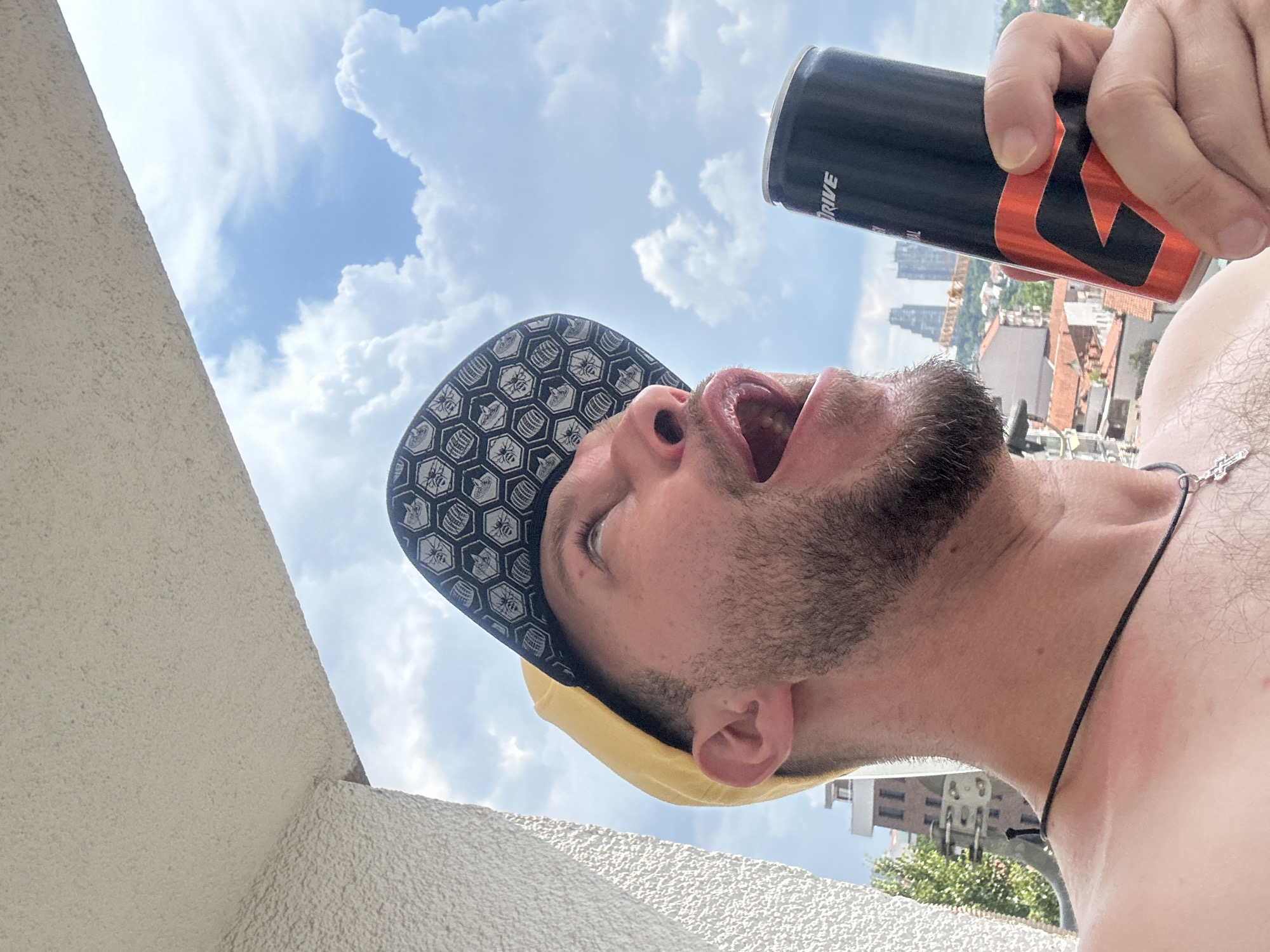 In the Zvezdara Urban Municipality of Serbia, a man wearing a yellow baseball cap and a black patterned cap is holding a red and black energy drink can. He has a beard and is looking upwards with his mouth open, as if about to drink from the can. The background shows a sky with white clouds and a city skyline with buildings and red-tiled roofs. The man is on a balcony, indicated by the textured wall on the left side of the image.