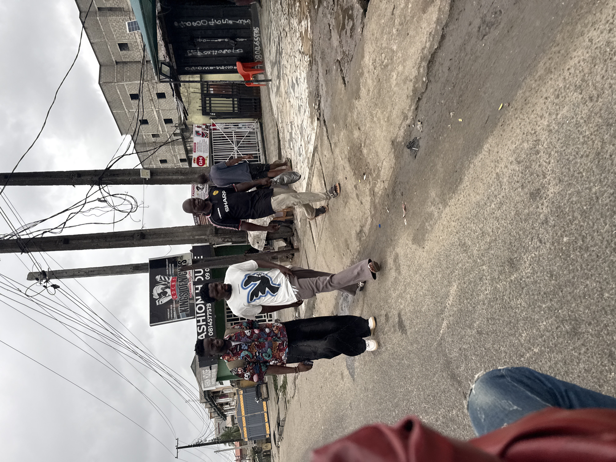 The image captures a street scene in Ado, Nigeria, under an overcast sky. Two young men are walking towards the viewer on a cracked asphalt road. The man on the left wears a colorful, patterned short-sleeved shirt, black baggy jeans, and white sneakers. He carries a backpack. The man on the right is dressed in a white t-shirt with a black graphic design and grey flared trousers, paired with sandals. Further in the background, another man in a black "SHARP" branded t-shirt and light-colored trousers walks away from the viewer, carrying a plastic bag. Behind him, a construction site is visible with unfinished brick buildings, and a bright orange chair stands out near a gated entrance. Several street signs, including one for "FASHION HOUSE" and another for "KINGSDEN'S CUTS," are present, indicating commercial activity. The environment appears somewhat neglected, with debris visible on the road and a general sense of urban grit.