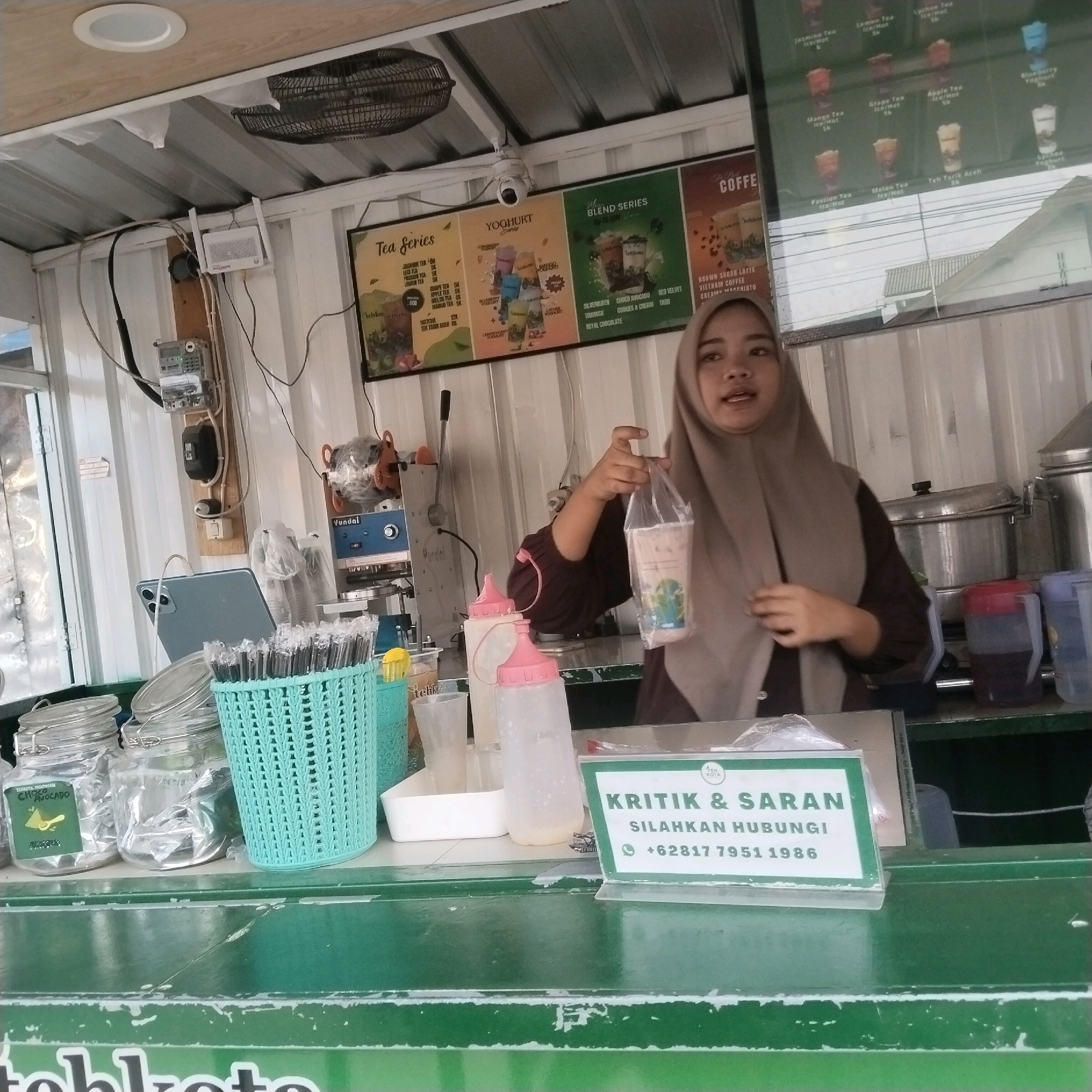 The image depicts an indoor scene at a beverage stall. A young woman, wearing a light brown hijab and a dark brown top, stands behind a counter, holding a clear plastic cup filled with a beverage and sealed with plastic. Her right hand is extended, presenting the drink. The stall features several menu boards on the wall behind the counter, advertising various tea, yogurt, and coffee drinks. On the counter, there are glass jars, a turquoise basket filled with straws, and plastic bottles. A tablet is propped up on the counter to the left. In the background, kitchen equipment, including a sealing machine and large metal containers, are visible. A sign in the foreground reads "KRITIK & SARAN SILAHKAN HUBUNGI +62817 7951 1986", indicating a customer feedback channel. The overall environment suggests a small business operating in Janteh, Indonesia, likely a food and beverage service establishment.