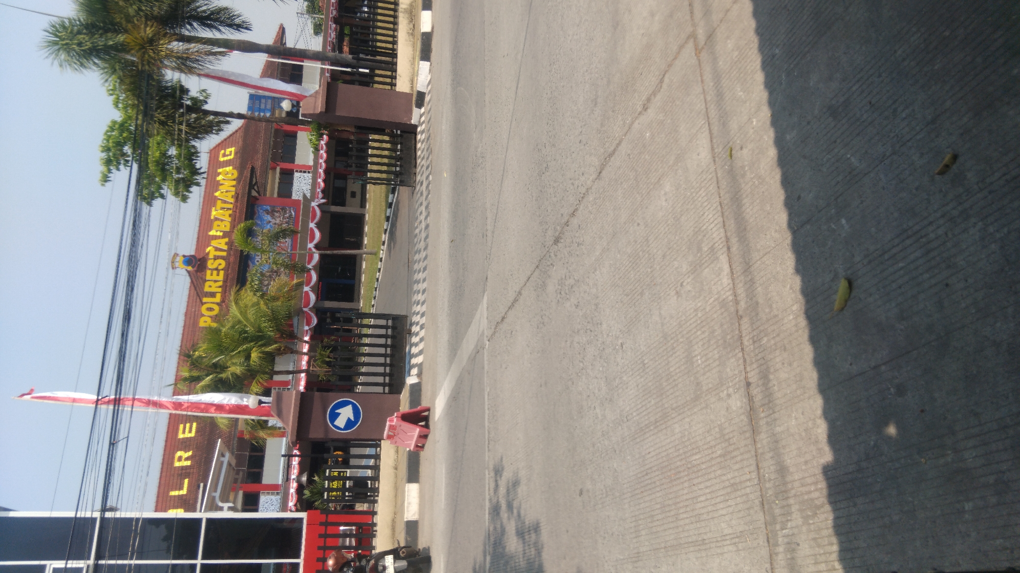 The image depicts the exterior of a building identified by a sign as "POLRESTA BATANG G". The building has a reddish-brown tiled roof and is painted in shades of red, white, and dark blue. There are large gates in front of the building, partially open, with a black metal fence. A red traffic barrier is positioned on the road in front of the gates. The road itself is paved with concrete and shows visible tire marks and lane markings. Palm trees and other foliage are present on the grounds of the building, and two flags, one red and white and another white, are flying from poles. The sky is clear and blue, indicating a sunny day. The overall scene suggests a public building, likely a police station, in a location that appears to be in Indonesia, given the signage.