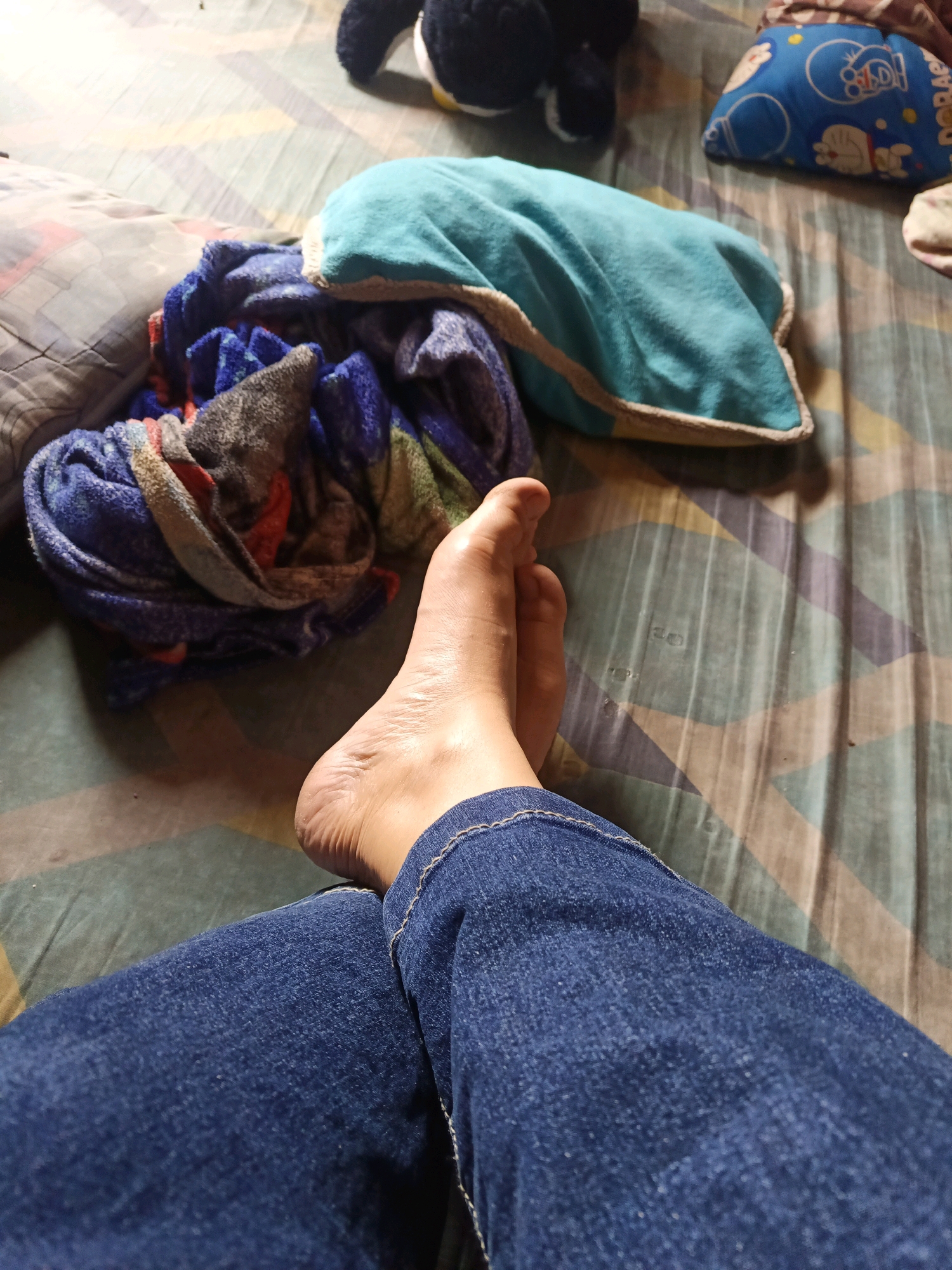 A close-up, high-angle shot shows a person's bare foot and lower legs, clad in blue denim jeans, resting on a striped bed in South Jakarta, Indonesia. The foot is positioned in the foreground, with the toes pointing upwards and slightly to the left. The skin is smooth with visible texture on the heel. The jeans are a dark blue wash with visible stitching. Behind the foot, a pile of folded blankets in shades of blue, gray, and red are present. A teal blanket with a cream-colored trim lies on top of the other blankets. In the background, to the right, a blue throw pillow with a cartoon character and lettering is visible. Further in the background, a portion of a dark plush toy, possibly a penguin, is seen. The bedsheets have a geometric pattern of green, beige, and purple stripes. The lighting suggests an indoor setting, possibly a bedroom, with a warm, soft glow.