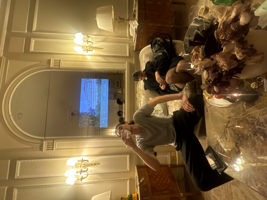 A luxurious living room with two people casually lounging on an ornate sofa. One person in a gray shirt drinks from a glass while the other reclines in a black jacket and shorts. A large marble coffee table is adorned with a large bouquet of flowers, plates, and various objects. A large mirror dominates the wall, reflecting a TV playing a video. Decorative wall lamps and ornate molding accentuate the room's opulence. A bottle of champagne sits on a table nearby.