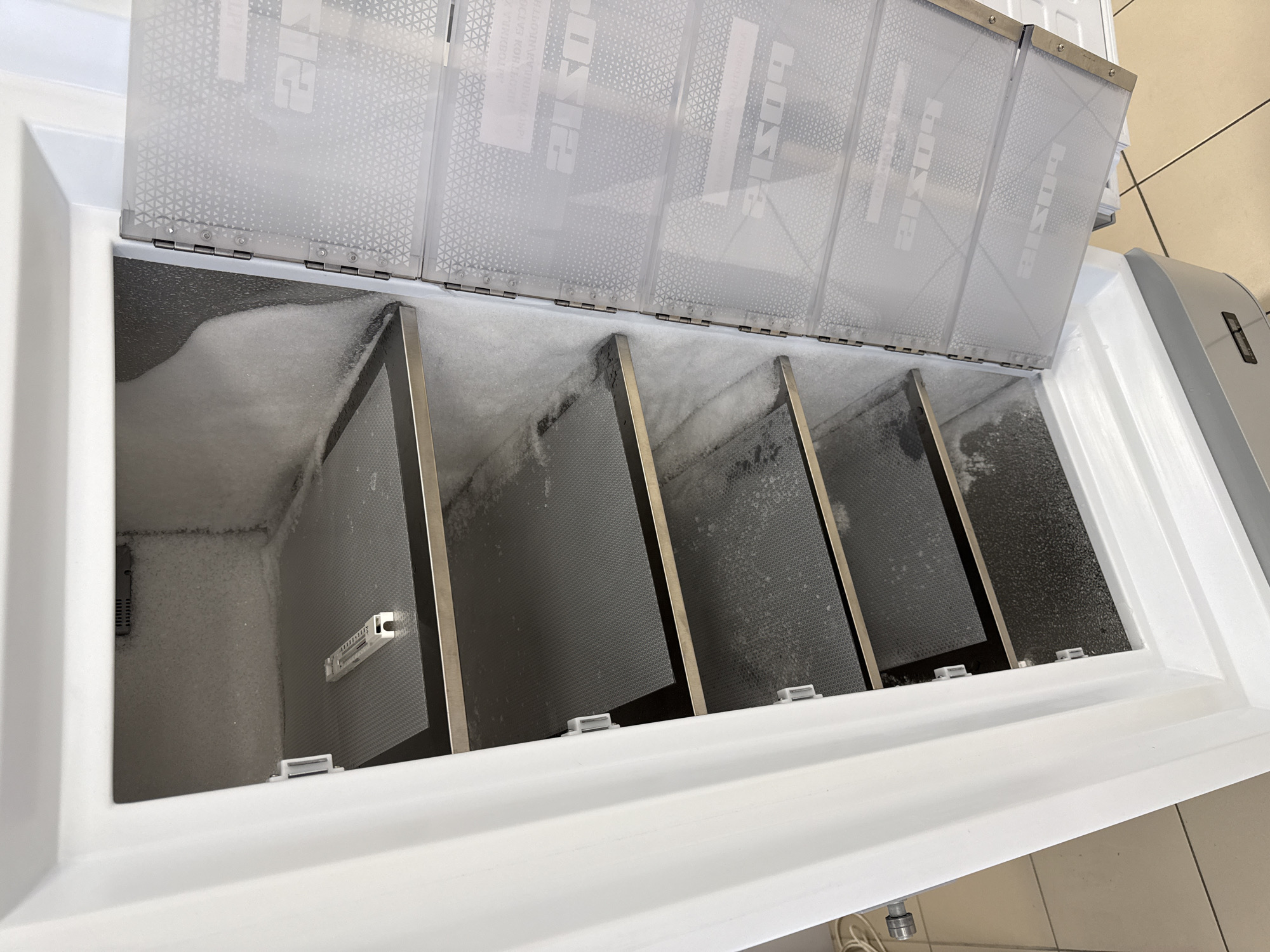 The image displays the open interior of a white chest freezer, likely a commercial unit, in an indoor setting in Moscow, Russia. Significant white frost and ice accumulate on the left internal wall and parts of the freezer's base, indicating continuous operation or a need for defrosting.
Above the main freezer compartment, several hinged, semi-transparent plastic panels form a secondary lid. These panels feature a subtle triangular mesh pattern and display the brand name "BOSIE" printed in reverse, along with other faint, reversed text, notably "INHALT KONTROLLE" (content control). Beneath these panels, four dark grey, perforated metal dividers with metallic frames are positioned upright, segmenting the freezer's internal space. A small, white, rectangular device, possibly a thermometer or sensor, is clipped to one of these dividers on the left side. Small white plastic clips along the freezer's front edge secure the dividers. The background shows a light-colored tiled floor, typical of a commercial or storage environment. No individuals are present in the scene.