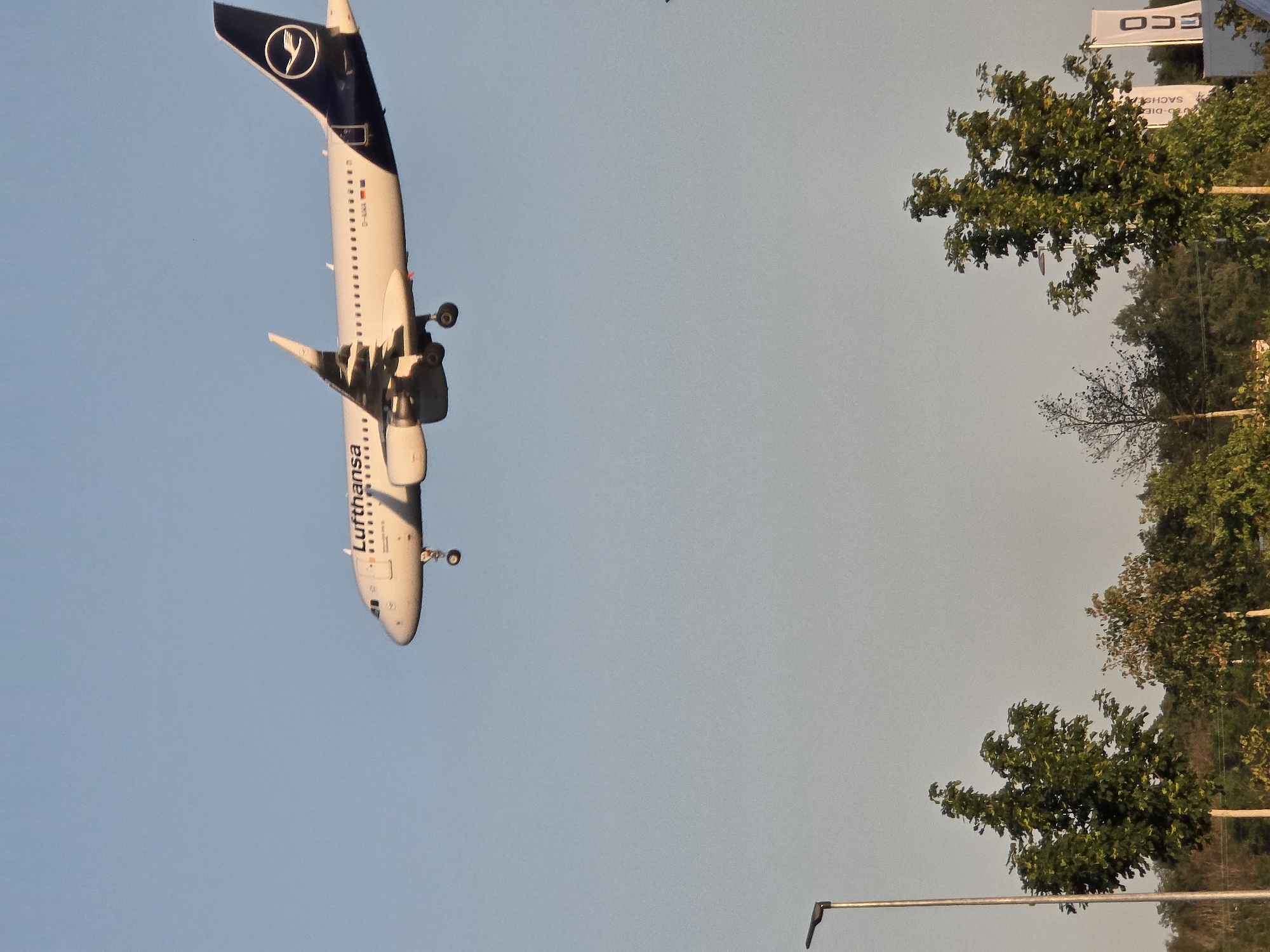 The media file shows a Lufthansa airplane in flight against a clear, pale blue sky. The plane is primarily white with a dark blue tail featuring the Lufthansa logo, which is a stylized crane. The word "Lufthansa" is visible on the fuselage in dark lettering. The landing gear is extended, suggesting the aircraft is either preparing for landing or has just taken off.
In the background, on the right side of the frame, are trees with lush green foliage, indicating a pleasant, likely warm, season. Above the trees, a white banner with black lettering is partially visible. The visible text on the banner appears to be "ECO" and potentially "SACHS", suggesting a location near an ecological or industrial zone, possibly related to an airport. The orientation of the image is unusual, with the airplane appearing to fly upside down, which could be due to camera tilt or rotation.
The lighting suggests it is daytime, with soft sunlight illuminating the scene. There are no people visible in the frame. The overall impression is of a typical operational scene at an airport or in its vicinity, with the focus being the airplane in transit. The presence of the trees and clear sky suggests an outdoor setting. Given the location context of Kelsterbach, Germany, this scene is likely near Frankfurt Airport.