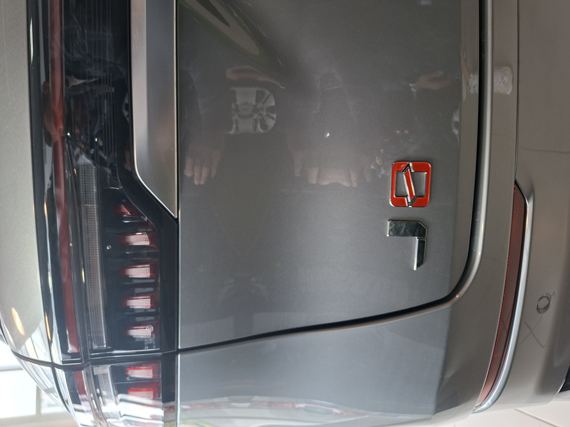 The image shows the rear of a grey car, likely a luxury sedan. The car has distinctive red and chrome taillights that span the width of the rear. Below the taillights, on the trunk, are two badges. The left badge is a chrome "J", and the right badge is a red and chrome "0". The car is parked indoors, possibly in a showroom or garage, with reflections of lights and people visible on its polished surface. One reflection shows a person with their hands out, as if taking a photo or admiring the car. The environment appears clean and well-lit, suggesting an organized space. There is no indication of the city of Izhevsk, Russia, in the image itself.