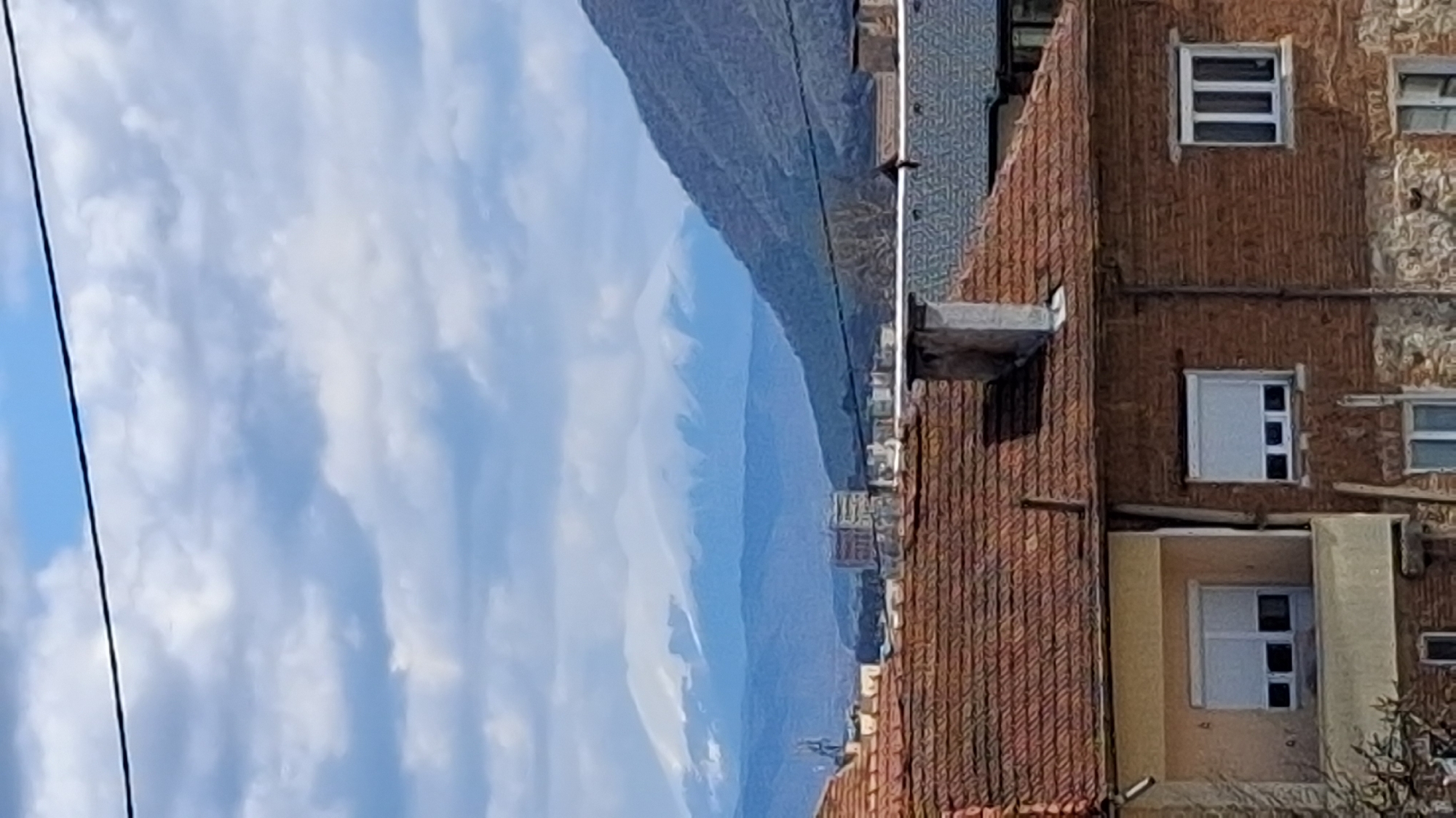 This image shows a vertical view of a scene in Goce Delchev, Bulgaria. The sky, filled with white clouds, occupies the left part of the image. Mountains are visible in the background. On the right, there's a building with a red-tiled roof and brick facade with several windows. A utility line runs along the top left portion of the frame.