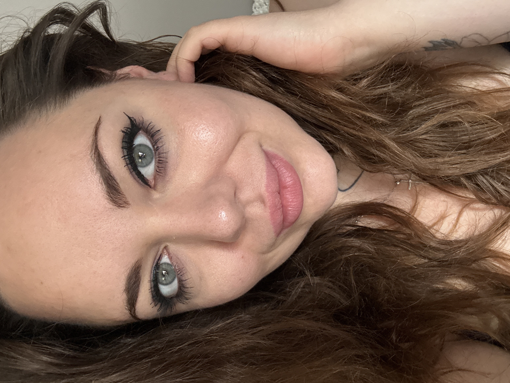 The image is a close-up portrait of a woman with light skin, brown hair, and blue eyes. She is wearing makeup, including eyeliner and mascara. Her lips are painted a light pink color. She has a subtle smile and her right hand is touching her ear. There is a faint tattoo visible on her neck. The background is out of focus, but appears to be an indoor setting. There is no information in the image to suggest a location in Kremenchuk, Ukraine.