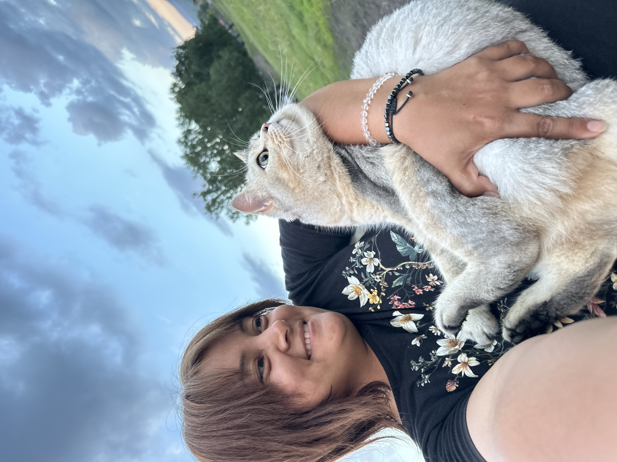 An outdoor, low-angle, selfie-style, medium shot captures a woman holding a light-colored cat against a cloudy sky. The woman, positioned on the left, has brown hair and is smiling. She is wearing a black t-shirt with a floral print. The cat, facing away from the camera and looking up, is held in the woman's arms. The background consists of a grassy hill with trees and a dramatic sky. The scene is set in an unspecified outdoor location, possibly a park or a rural area, with no identifiable city landmarks.