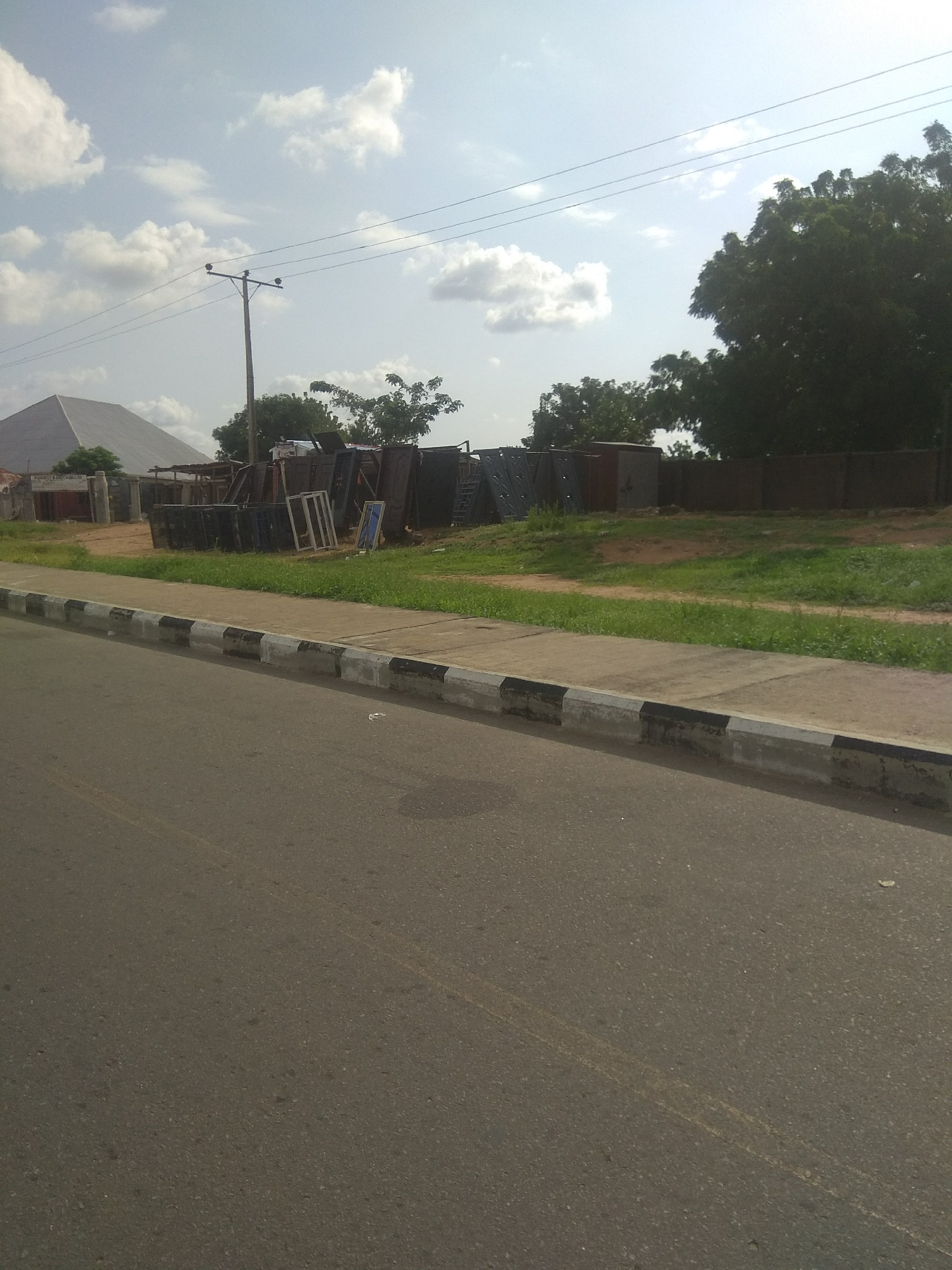 The image shows a street scene in Jalingo, Nigeria, with a paved road in the foreground and a sidewalk bordering a grassy area. Behind the sidewalk, a collection of various items, including what appear to be metal window frames and possibly building materials, are stacked against a fence. A large, pyramidal-shaped structure, possibly a building or a roofing project, is visible in the background to the left, with a utility pole and power lines extending across the sky. Lush green trees line the right side of the scene, with a cloudy sky overhead. There are no people or vehicles visible in the image.