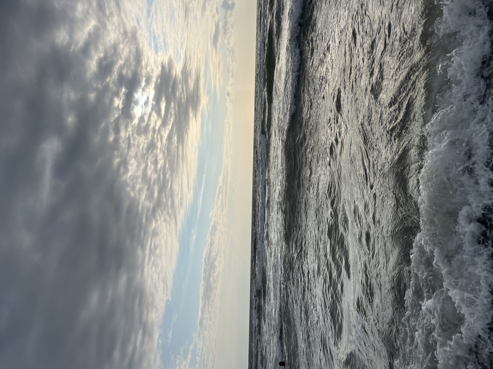 The image shows a view of the Black Sea from the shore at Ureki, Georgia. The sky is partly cloudy, with patches of sunlight breaking through the gray clouds, creating a dramatic effect. The sea is choppy, with waves rolling towards the shore. The foreground shows white foam from the breaking waves. In the distance, a person can be seen swimming in the sea, on the left side of the image. The horizon line is visible, separating the sea from the sky. The overall mood of the image is serene yet powerful, capturing the dynamic nature of the ocean under a cloudy sky.