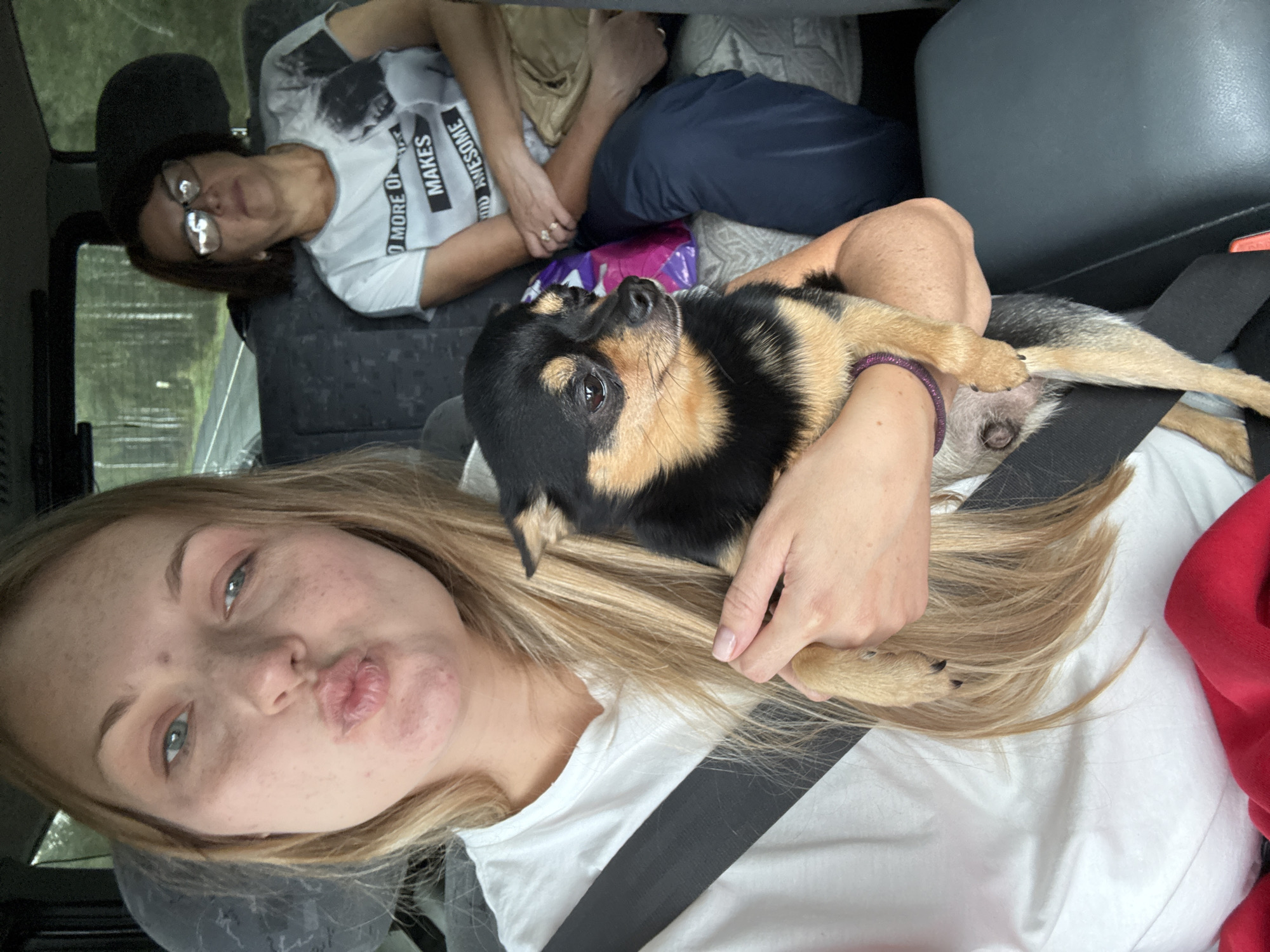 In the city of Ryzhkovo, Russia, a woman is seated in the passenger seat of a vehicle, holding a small black and tan dog in her arms. She is looking at the camera with her lips pursed. She is wearing a white t-shirt and a seatbelt. In the back seat behind her, another woman is seated, wearing glasses and a t-shirt with a graphic print. She is looking away from the camera. The interior of the vehicle appears to be a van or SUV, with dark grey upholstered seats visible. Outside the vehicle, a wooded area can be seen through the windows, suggesting the vehicle is in a rural or natural setting. The lighting indicates it is daytime.