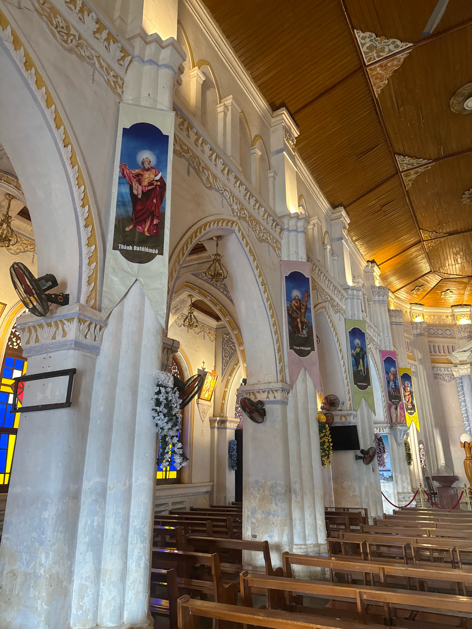 The image captures the interior of a Roman Catholic church in Hội Tín, Vietnam. The view is from a low angle, looking up towards the ceiling and along the nave. White, fluted columns, adorned with decorative gold trim, line the sides of the church. Suspended from the columns are several large, rectangular banners depicting religious figures, each with text in Vietnamese.
The ceiling is constructed of wooden planks and beams, with decorative corner accents. Multiple electric fans are mounted on the columns, indicating a warm climate. Stained glass windows with vibrant blue and yellow panes are visible on the left side of the frame. Rows of empty wooden pews fill the foreground and extend down the nave, suggesting the church is either empty or the congregation is not yet present or has dispersed. The lighting appears to be artificial, with warm light illuminating the architectural features and banners. The overall impression is one of reverence and historical architecture.