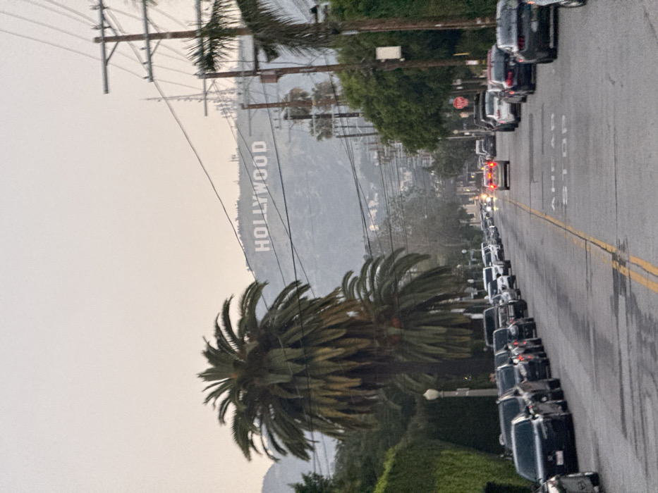 This image depicts a residential street scene in the city of Los Angeles, United States of America, specifically oriented towards the Hollywood Hills.
In the background, the iconic "HOLLYWOOD" sign, composed of large white capital letters, is prominently visible on a distant, brush-covered hillside. Multiple communication towers and structures are also present on the hill. The sky is overcast or hazy, obscuring the horizon.
The midground features a straight, two-lane asphalt street with numerous vehicles parked bumper-to-bumper along both curbs. These vehicles are primarily sedans and SUVs of various colors. On the left side of the street, several mature palm trees with dense, green fronds stand tall, partially obscuring the view of adjacent properties, which include a manicured green hedge and a classic street lamp post. On the right side, utility poles with multiple strands of overhead power lines extend vertically and horizontally across the frame. One light-colored sedan with illuminated red brake lights is observed driving away from the viewer in the right lane of the street. A red octagonal stop sign is partially visible on the right side of the road, further down the street.
In the foreground, the street surface displays a faded double yellow center line and the painted white text "AHEAD STOP" on the asphalt, indicating an upcoming traffic stop. No individuals are visible in the scene. The lighting suggests dusk or an overcast daytime condition.