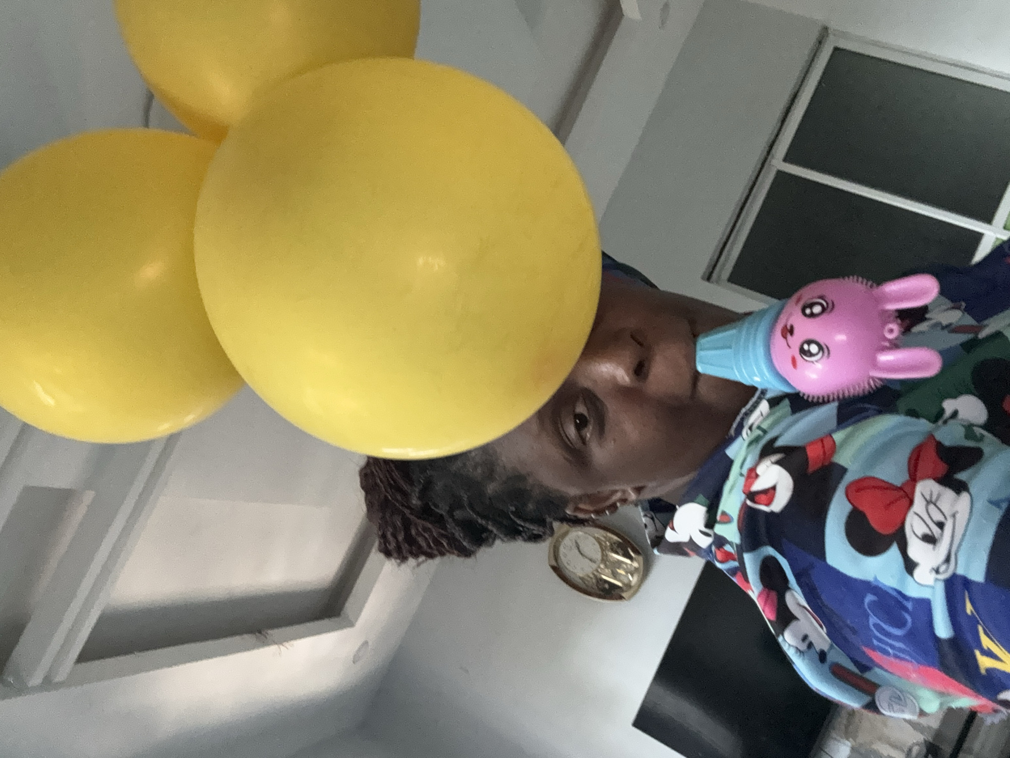 A person is visible from the chest up, with yellow balloons partially obscuring their face. They are wearing a blue and white shirt with Disney characters. To the person's right is a pink and blue bunny-shaped toy. The background includes a window with horizontal bars and what appears to be the interior of a room with white walls. Based on the prompt, this scene takes place in Sabon Tasha, Nigeria, but there are no visual cues to confirm the location.