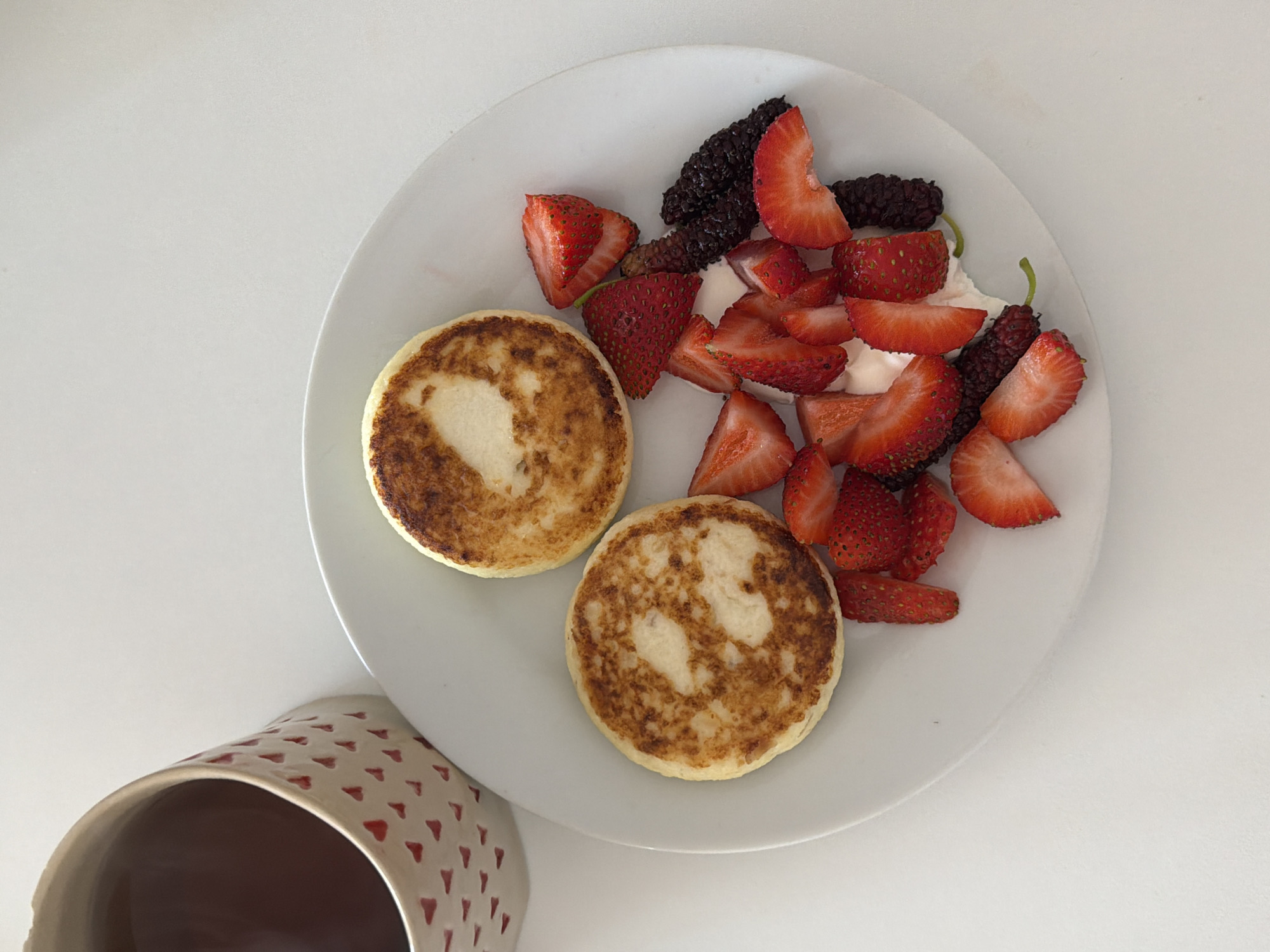 A high-angle, brightly lit studio shot features a white plate containing two golden-brown, round cottage cheese pancakes (syrniki) accompanied by cut strawberries and mulberries, with a dollop of white cream. Beside the plate is a portion of a cream-colored mug with small red heart designs, filled with a dark liquid, possibly coffee or tea. The background is a plain white surface. The scene is a domestic setting. The location in the city of Tel-Aviv, Israel, cannot be determined from the image.