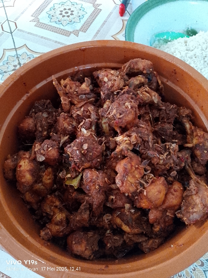 The image shows a close-up of a large brown ceramic bowl filled with a dark, rich, stewed chicken dish, likely a traditional Indonesian recipe. The chicken pieces appear tender and fall-off-the-bone, with a dark, caramelized sauce coating them. There are visible pieces of fried onion on top of the chicken, adding a contrasting texture and flavor. The dish appears to be freshly prepared and very appetizing. It is not possible to confirm the exact location in Gegesik, Indonesia, using only the coordinates provided. The image was likely taken indoors based on the visible patterned floor tiles and the bowl's placement on a table.