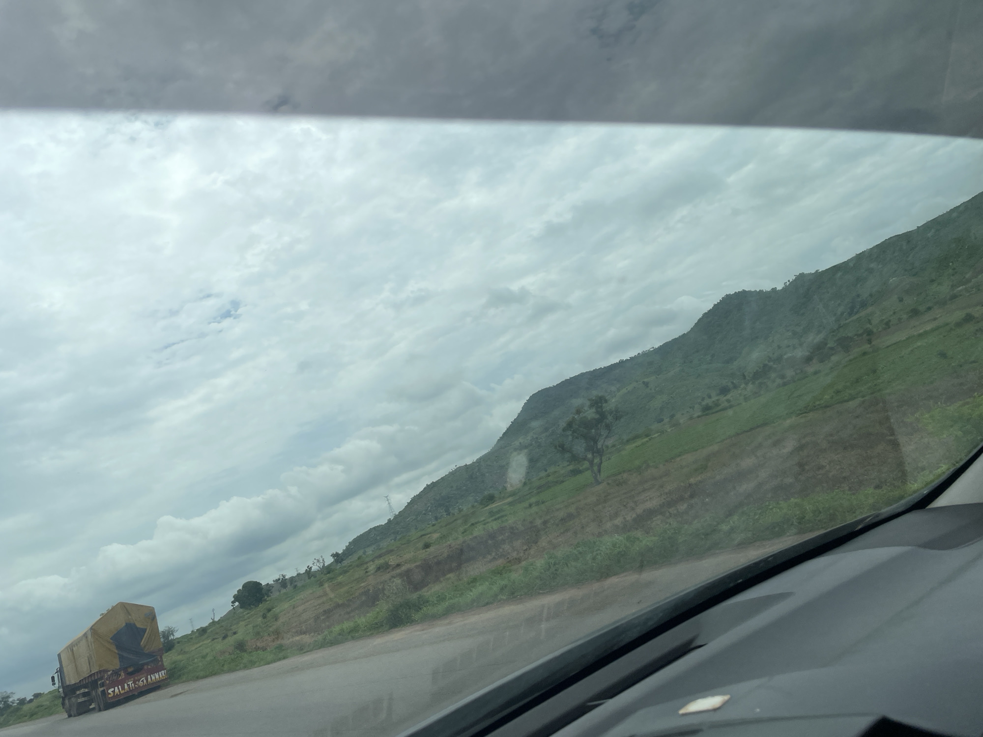This media file depicts a scenic view from inside a vehicle, likely a car, traveling on a road in Kwali, Nigeria. The scene is dominated by a vast, cloudy sky and a lush green hillside. The road itself is visible in the foreground, with the dashboard of the vehicle partially obscuring the lower portion of the frame.
The hillside is terraced, suggesting agricultural activity. Patches of cultivated land and vegetation are interspersed across its slopes. A lone tree stands prominently on the hillside. In the distance, a large truck covered with a yellow tarp is driving on the road, its side displaying some text. The sky is filled with a mix of light and dark clouds, indicating overcast or potentially stormy weather. The overall impression is one of a journey through a rural, hilly landscape under a dramatic sky. The time of day appears to be daytime, possibly late morning or afternoon.
The text visible on the truck reads "SALAT&ANNARI".