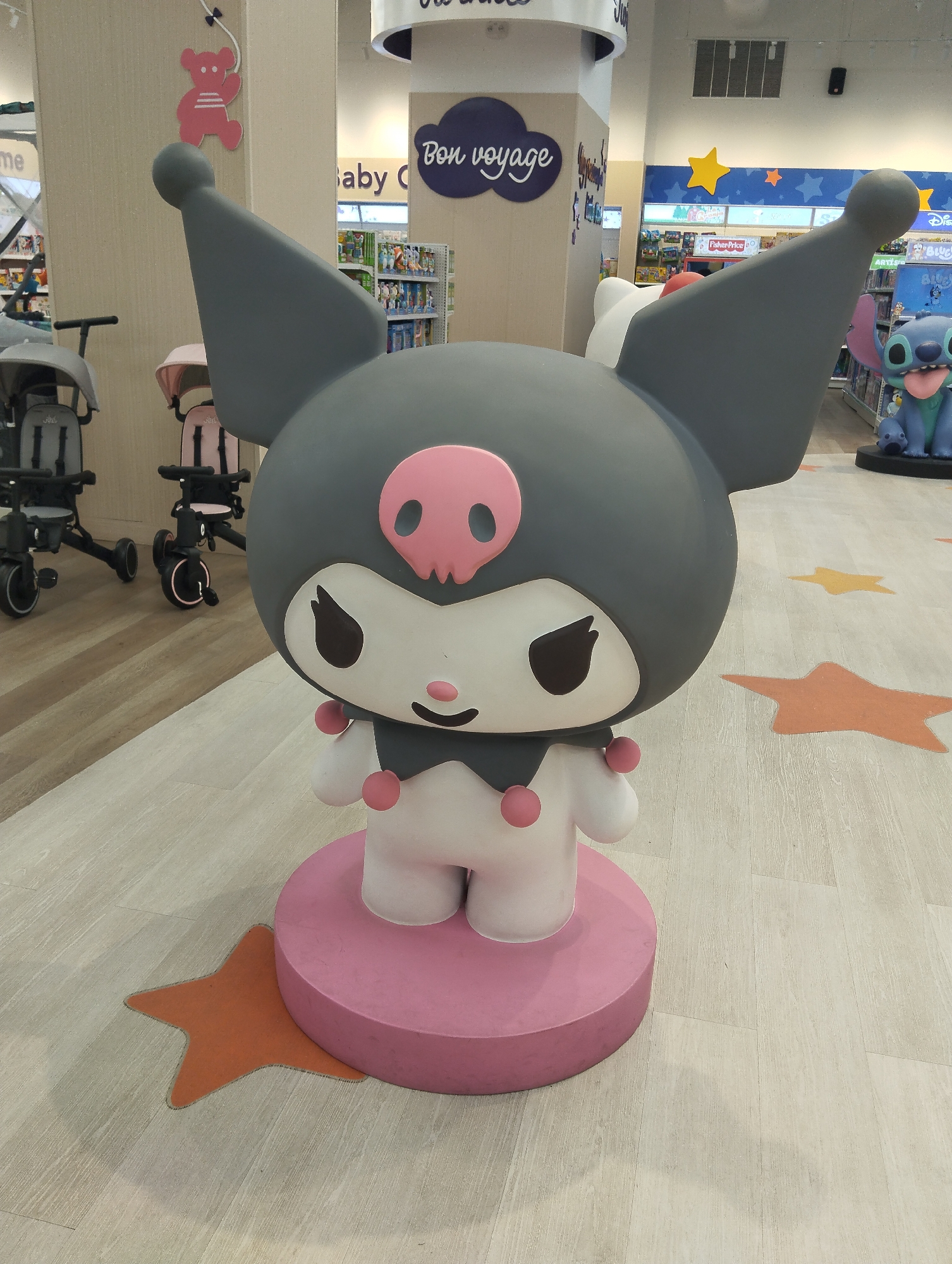 A large statue of Kuromi, a character from the Sanrio universe, dominates the foreground of this indoor retail scene in Porlamar, Venezuela. The Kuromi statue is predominantly dark gray and white, with prominent pink details including a skull emblem on its forehead, pink cheek dots, and a round pink base. Its large, pointed ears and mischievous expression are clearly visible.
The setting appears to be a brightly lit children's section of a department store or a toy store. The floor is a light-colored wood-effect laminate, decorated with various orange and yellow star-shaped cutouts.
In the background, several items related to children's products are visible. To the left, two strollers or tricycles, one dark gray and one pink, are on display. Behind them, a pink bear silhouette hangs as a decoration on a light-colored pillar. Further back, shelves filled with assorted merchandise are visible, with sections featuring blue walls adorned with yellow star decorations. On the far right, a partial view of a large blue Stitch character statue (from Disney's Lilo & Stitch) can be seen next to shelves.
Visible text includes "Bon voyage" and "Baby" on overhead signage, suggesting departments for infants or travel-related items. Brand names such as "Fisher-Price" and "Disney" are also faintly discernible on product displays in the background. The scene conveys a cheerful, consumer-oriented environment designed for families with young children.