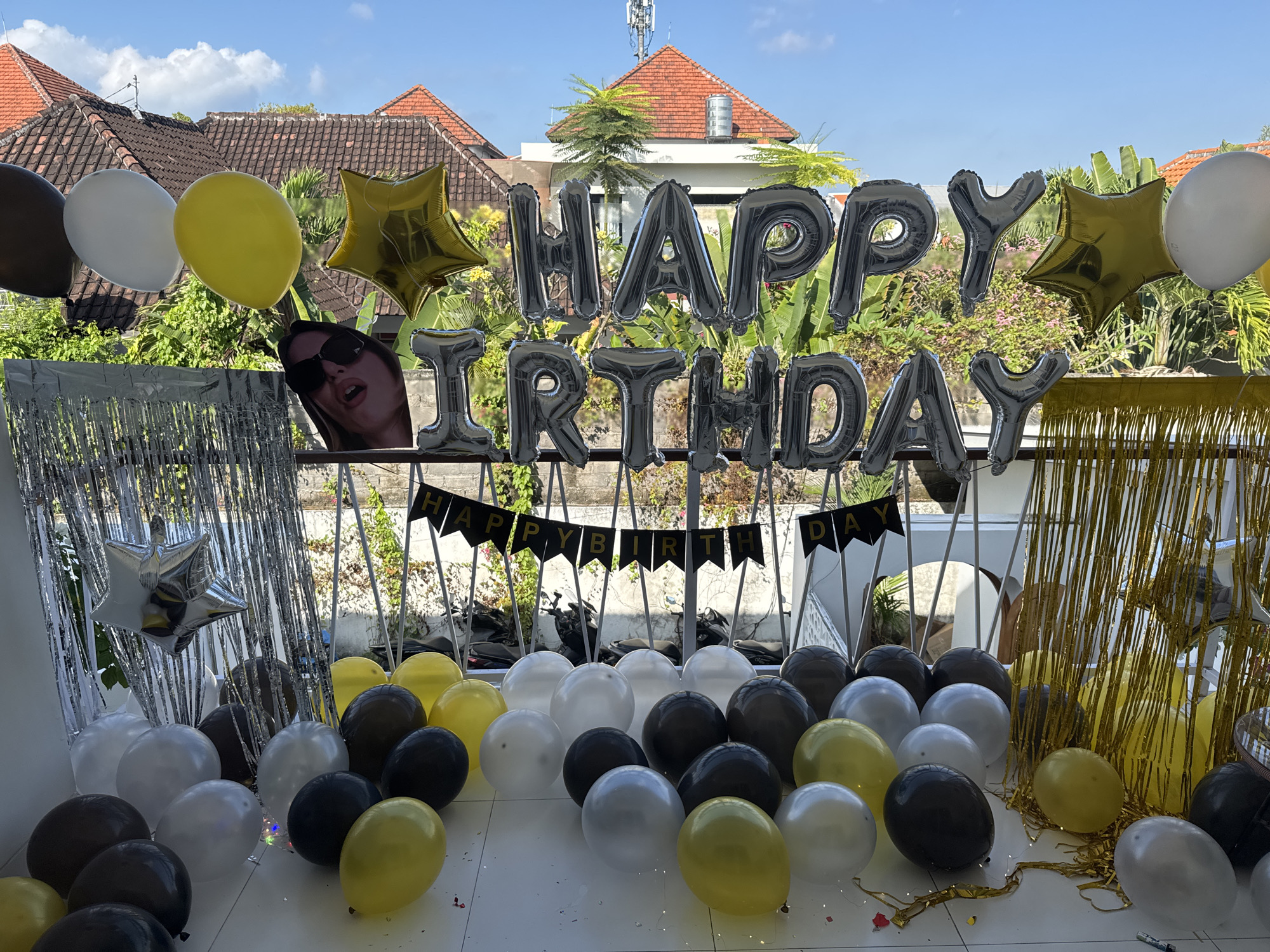 In North Kuta, Indonesia, a rooftop balcony is decorated for a birthday celebration. A collection of black, white, and yellow balloons are scattered across the floor and suspended in the air, along with foil balloons forming the words "HAPPY BIRTHDAY." A foil star balloon and a cardboard cutout of a woman with sunglasses are also present. Silver and gold fringe curtains hang on either side of the banner, adding a festive touch to the scene. The background reveals a residential area with tiled roofs and lush greenery, under a clear blue sky.