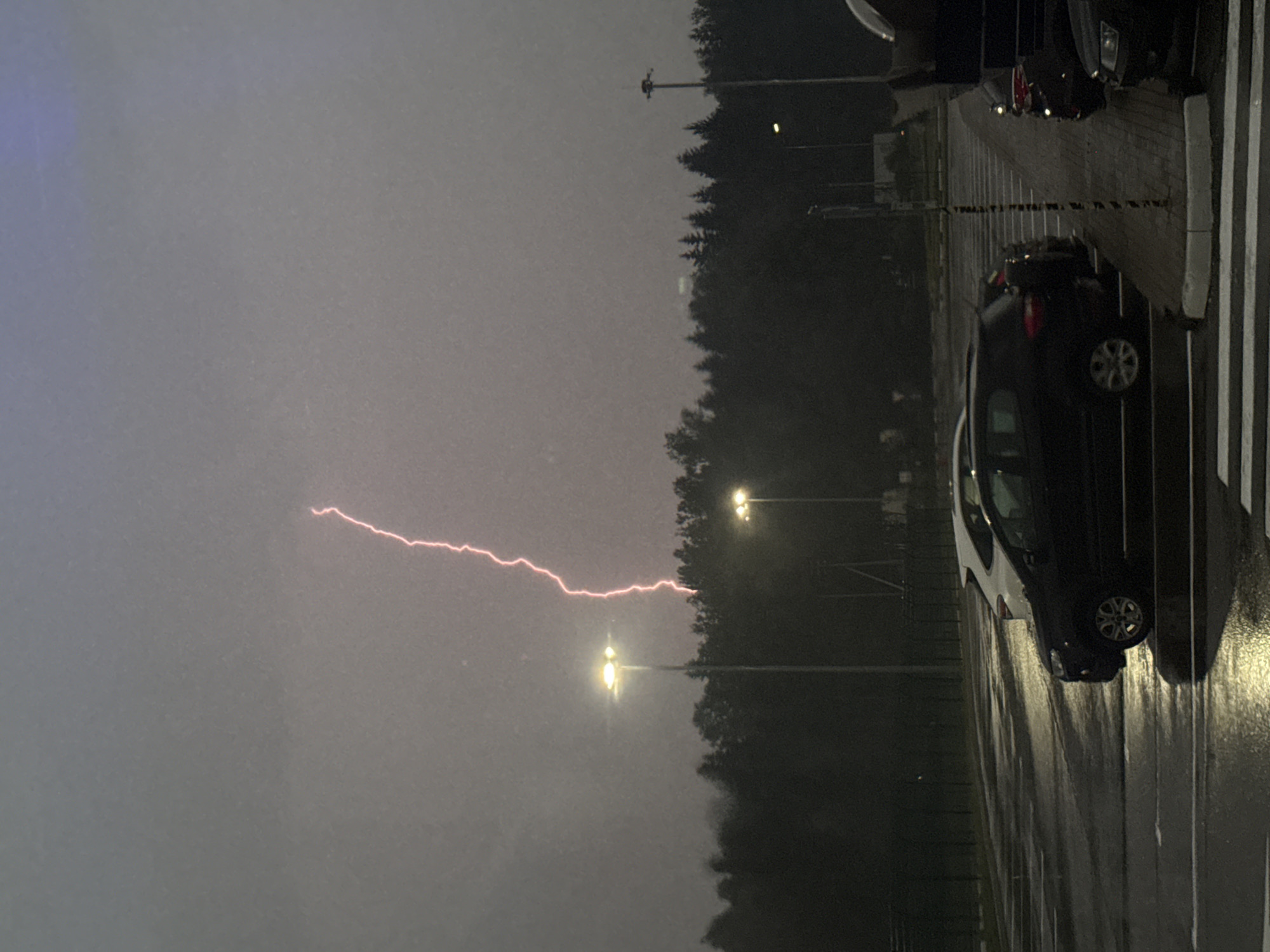 A lightning strike illuminates the sky over a tree line behind a parking lot. The sky is overcast and appears to be a dusky purple hue. Two lampposts are visible in the foreground, their lights on. A dark-colored SUV is parked in the foreground, and a white car is parked behind it. The ground in the parking lot is wet and reflective, suggesting it has recently rained. In the distance, a line of trees forms a dark silhouette against the sky.