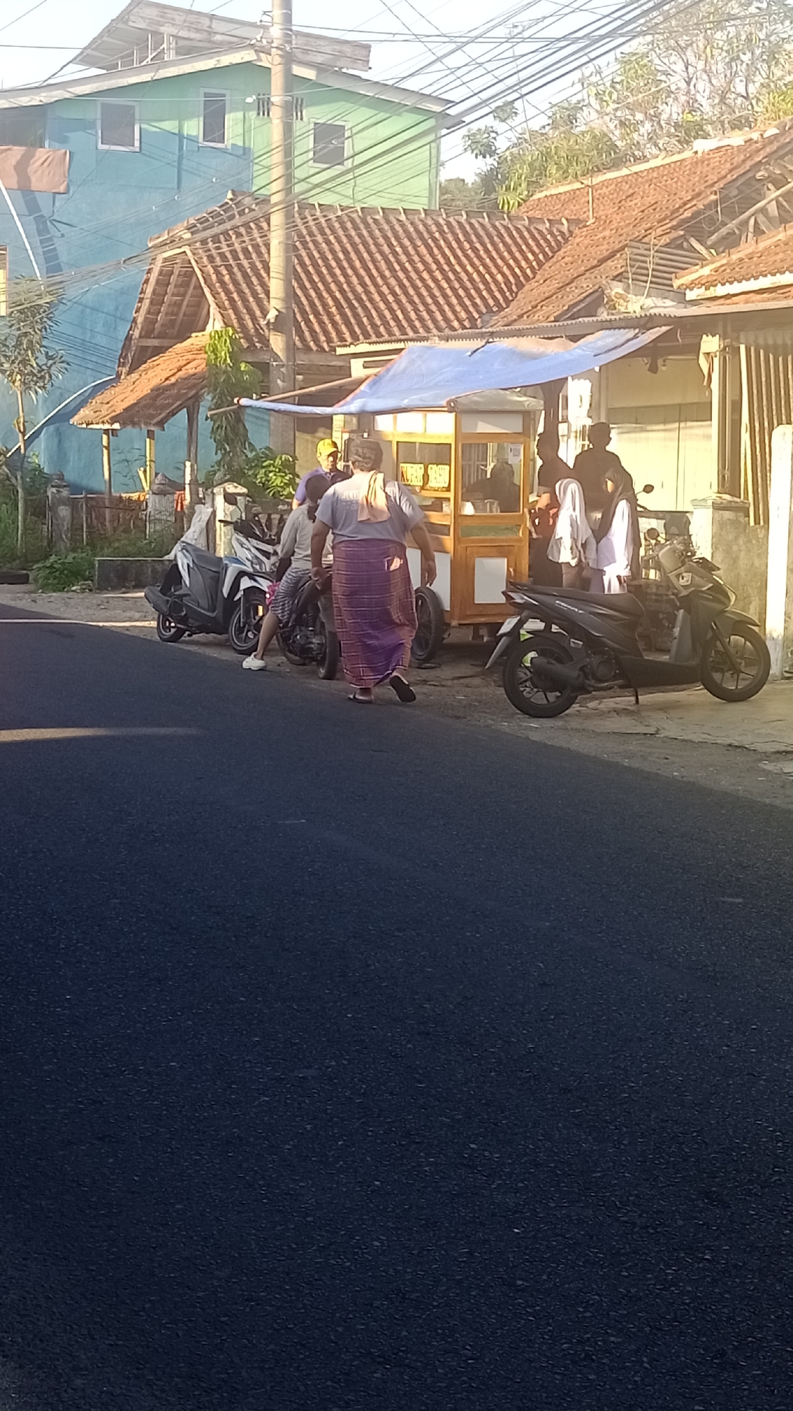 In Pakemitan, Indonesia, a street scene unfolds under the warm glow of the sun, with a roadside food stall serving as a focal point. The stall, a modest wooden structure topped with a blue tarp, is frequented by several individuals. One person, wearing a purple sarong and a grey t-shirt, walks away from the stall. Another, in a grey t-shirt and patterned shorts, is seated on a motorbike. A third person, wearing a yellow cap and blue shirt, stands near the stall, seemingly engaged in a conversation with someone inside. Two other individuals are partially visible near the stall, their interactions unclear. Several motorbikes are parked nearby, indicative of local transportation. The backdrop features buildings with reddish-brown tiled roofs and a light green structure, characteristic of Indonesian architecture, alongside a utility pole adorned with numerous electrical wires. The foreground is dominated by the dark asphalt of a street, suggesting a public thoroughfare.