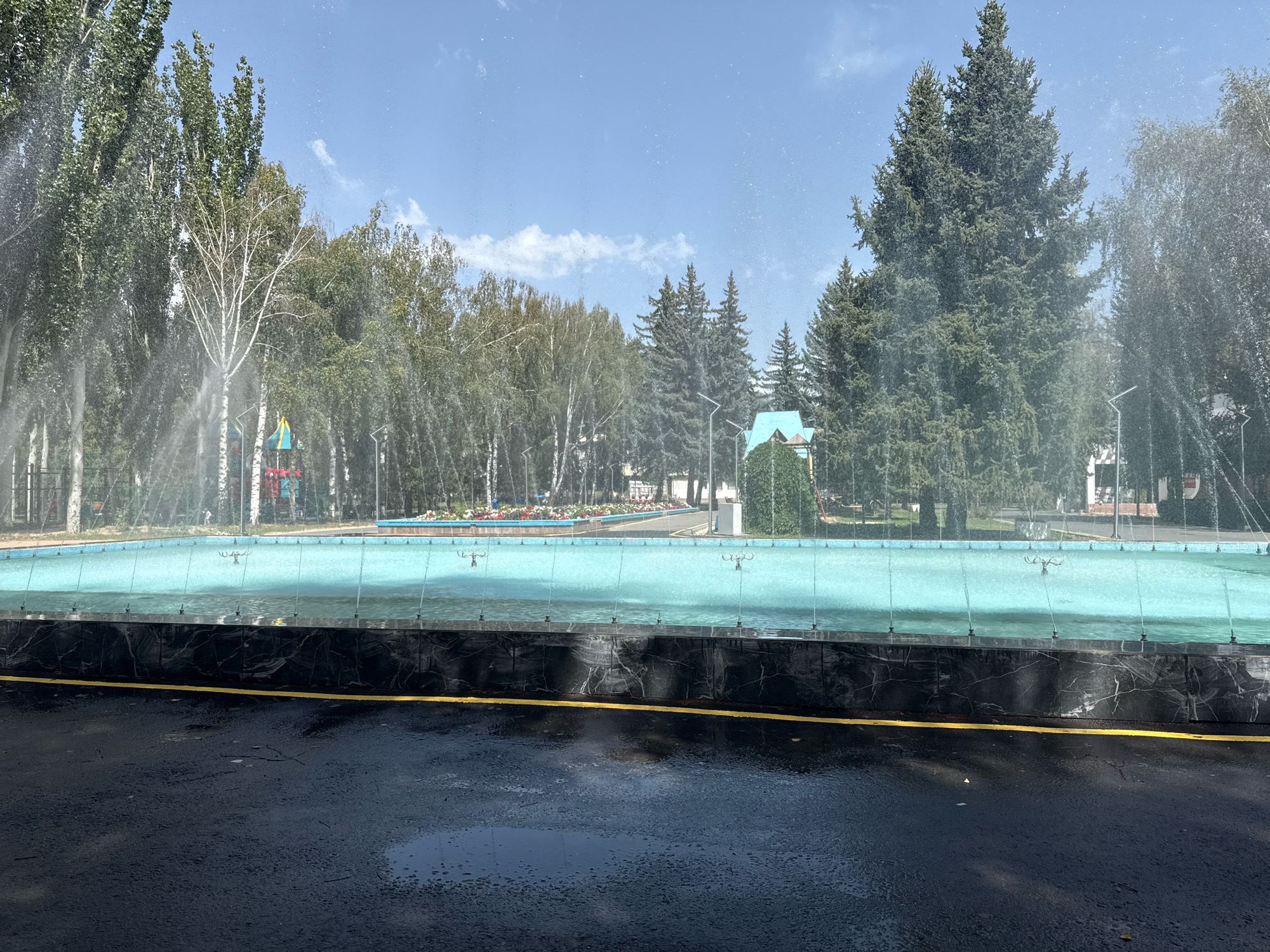 The image shows an empty, modern outdoor swimming pool filled with clear blue water. The pool is rectangular and surrounded by a dark, possibly marble, decorative border with a bright yellow line painted on the asphalt in front of it. In the background, tall trees line a park-like setting with a clear blue sky overhead. Spritzes of water are visible in the air, suggesting the fountain is active, and mist is creating a light haze over the scene. The asphalt in the foreground is wet and appears to have some puddles. The overall scene depicts a tranquil and well-maintained public space in Bosteri, Kyrgyzstan, possibly a resort area or park, on a sunny day.