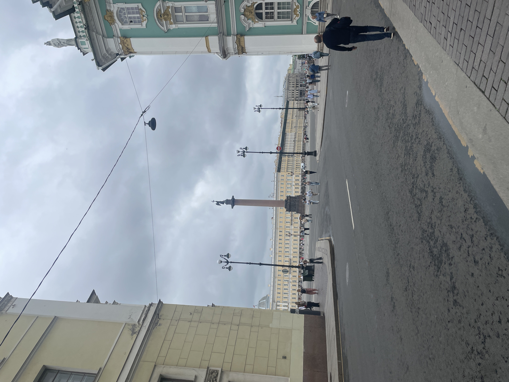 The image depicts a view of Palace Square in Saint Petersburg, Russia, under a cloudy sky. The foreground shows a paved street with a man in a dark jacket and jeans walking away from the camera on the right side. The left side of the street features a light yellow building with classical architectural elements. In the middle ground, a large square is populated with a number of pedestrians walking across the open space. Dominating the center of the square is the Alexander Column, a tall granite monument topped with a statue of the angel Michael. Behind the column, a long, ornate building with numerous windows stretches across the horizon, appearing to be the General Staff Building. On the right side of the image, a section of a building with turquoise and white facades, adorned with gold details and sculptures, is visible, likely part of the Winter Palace complex. Street lamps are present in the scene, and power lines are strung across the sky. The overall atmosphere is overcast and cool, suggesting a temperate climate.