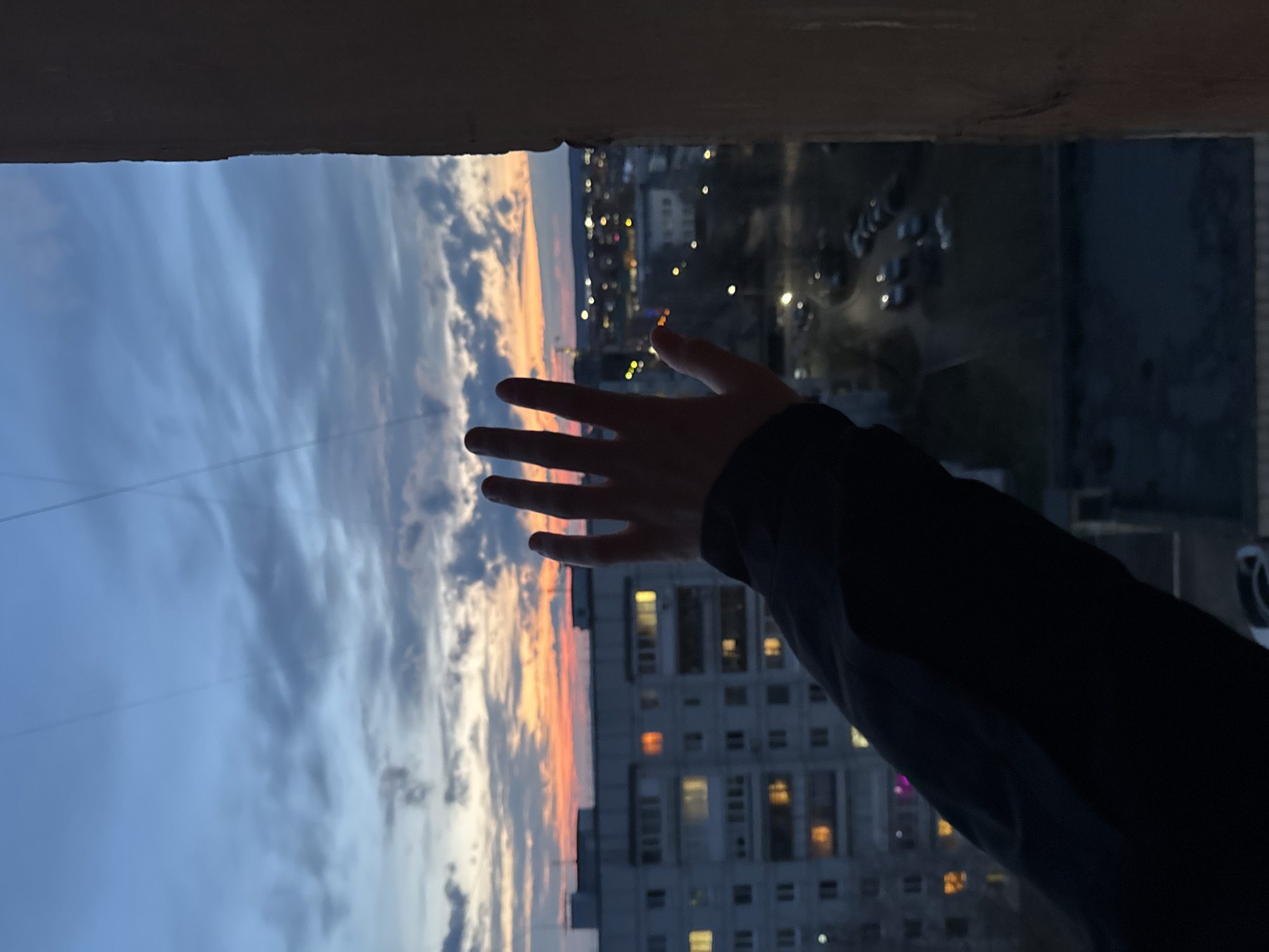 The image presents a first-person perspective from an elevated vantage point, likely a window or balcony, overlooking the city of Chaikovsky, Russia, during twilight. A human arm, clothed in a dark sleeve, stretches into the frame from the right, with an open hand and slightly spread fingers reaching towards the vast sky and urban expanse.
The upper and left portion of the frame is dominated by a dramatic sky, showcasing the vibrant hues of a fading sunset. Streaks of orange and pink clouds blend into softer blues and grays, indicating the transition from day to night. Below the horizon, the urban landscape begins to glow with artificial light. Numerous city lights appear as blurred bokeh in the mid-ground, suggesting the bustling activity of the city.
A prominent feature in the lower middle and right side of the image is a large, multi-story building, characteristic of post-Soviet architecture, with many windows, some brightly illuminated from within. Further to the right, a parking area is visible, dotted with vehicles and illuminated by streetlights. The scene captures the beauty of dusk over an urban setting. The outstretched hand adds a personal and contemplative element, creating a sense of connection or interaction with the expansive view. The overall mood is one of serene observation as evening descends upon the city. No visible text is present in the media.