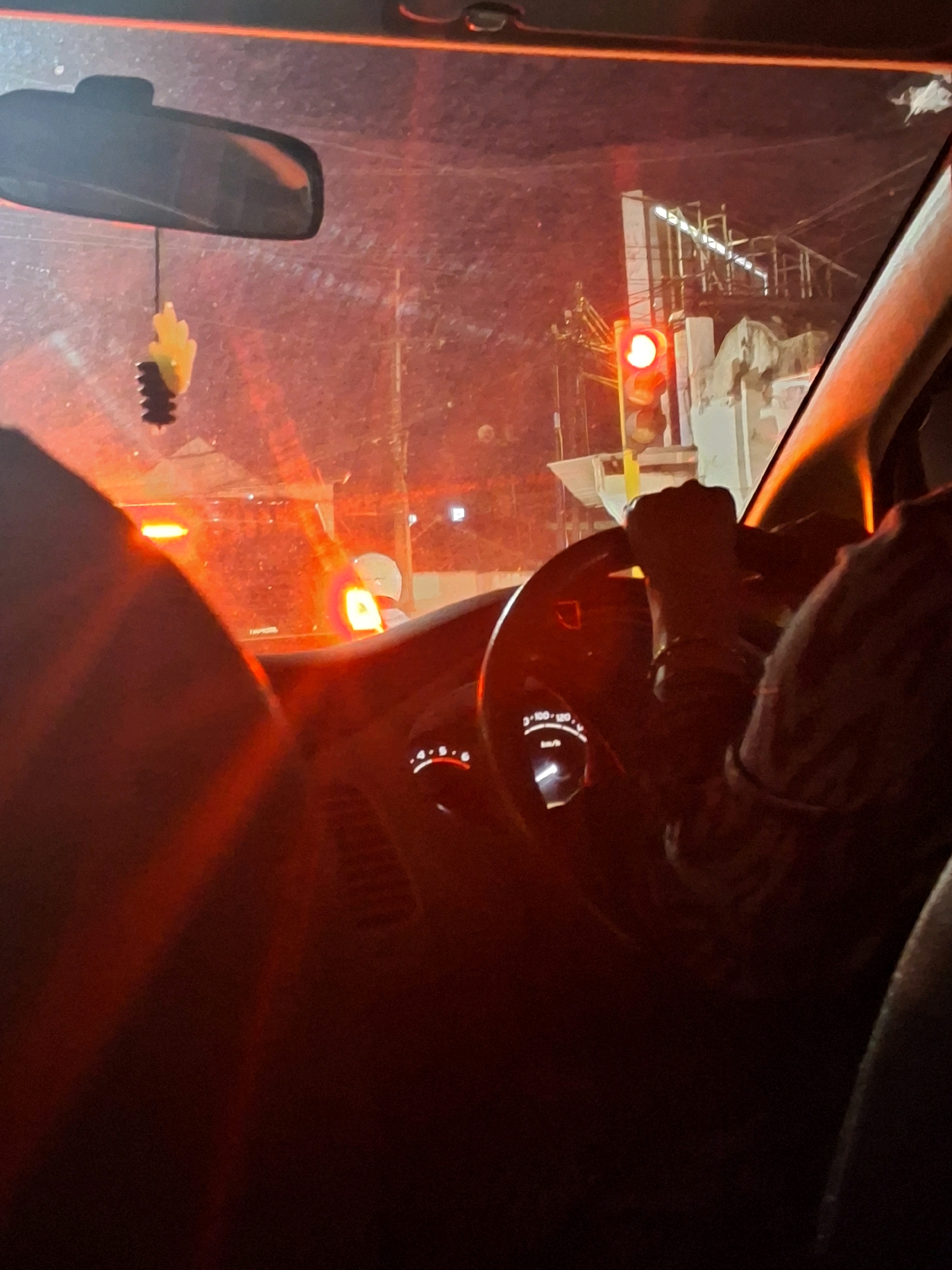 The image captures the interior of a car at night, likely in Lumajang, Indonesia, given the context. The scene is dominated by red light, originating from a traffic signal. A driver's hands are visible on the steering wheel, suggesting they are operating the vehicle. In the foreground, parts of the car's interior, including the dashboard with illuminated gauges, are discernible. Outside the car, a traffic light is prominently displayed, showing a red signal. The surrounding environment appears to be urban, with hints of buildings and infrastructure, possibly under construction, visible through the windshield. A car air freshener hangs from the rearview mirror. The overall impression is of a momentary pause at a traffic intersection in a city at night, illuminated by artificial lights.