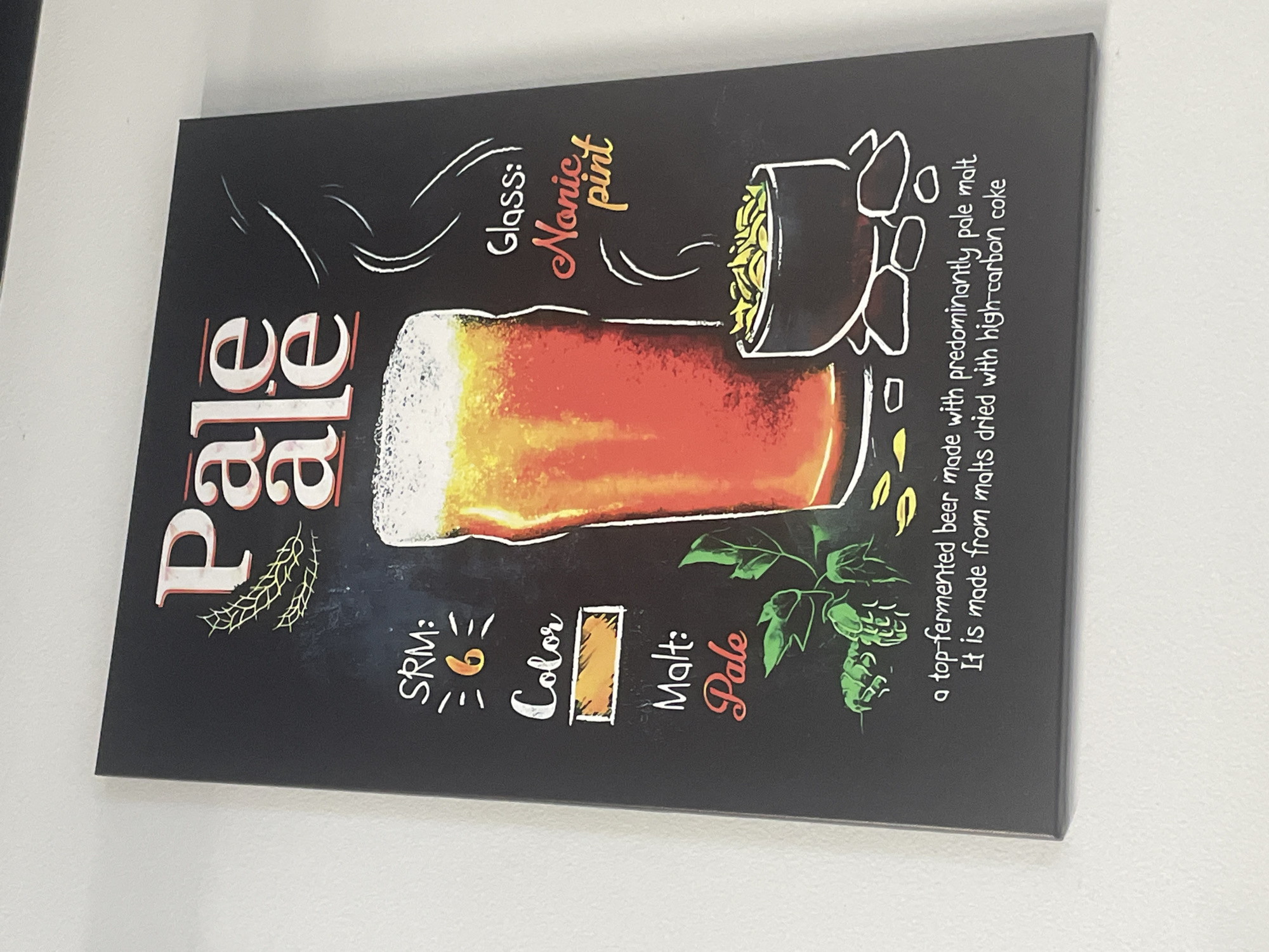 This is a photograph of a chalk-art style informational poster about "Pale Ale" displayed on a white wall. The poster has a black background and features white and colored text and illustrations. At the top, the words "Pale Ale" are written in large, stylized white lettering with red outlines. Below this, to the left, is information about the beer, including "SRM: 6", "Color" with a swatch of orange, and "Malt: Pale" with "Pale" in red script. To the right of these details is a drawing of a pint glass filled with amber-colored beer and a frothy head. Below the glass, a small black bowl contains yellow-green snacks, and stylized white lines indicate steam rising from the bowl. At the bottom of the poster, a description reads: "a top-fermented beer made with predominantly pale malt. It is made from malts dried with high-carbon coke." The poster is hung slightly askew, and the lighting casts a subtle shadow.